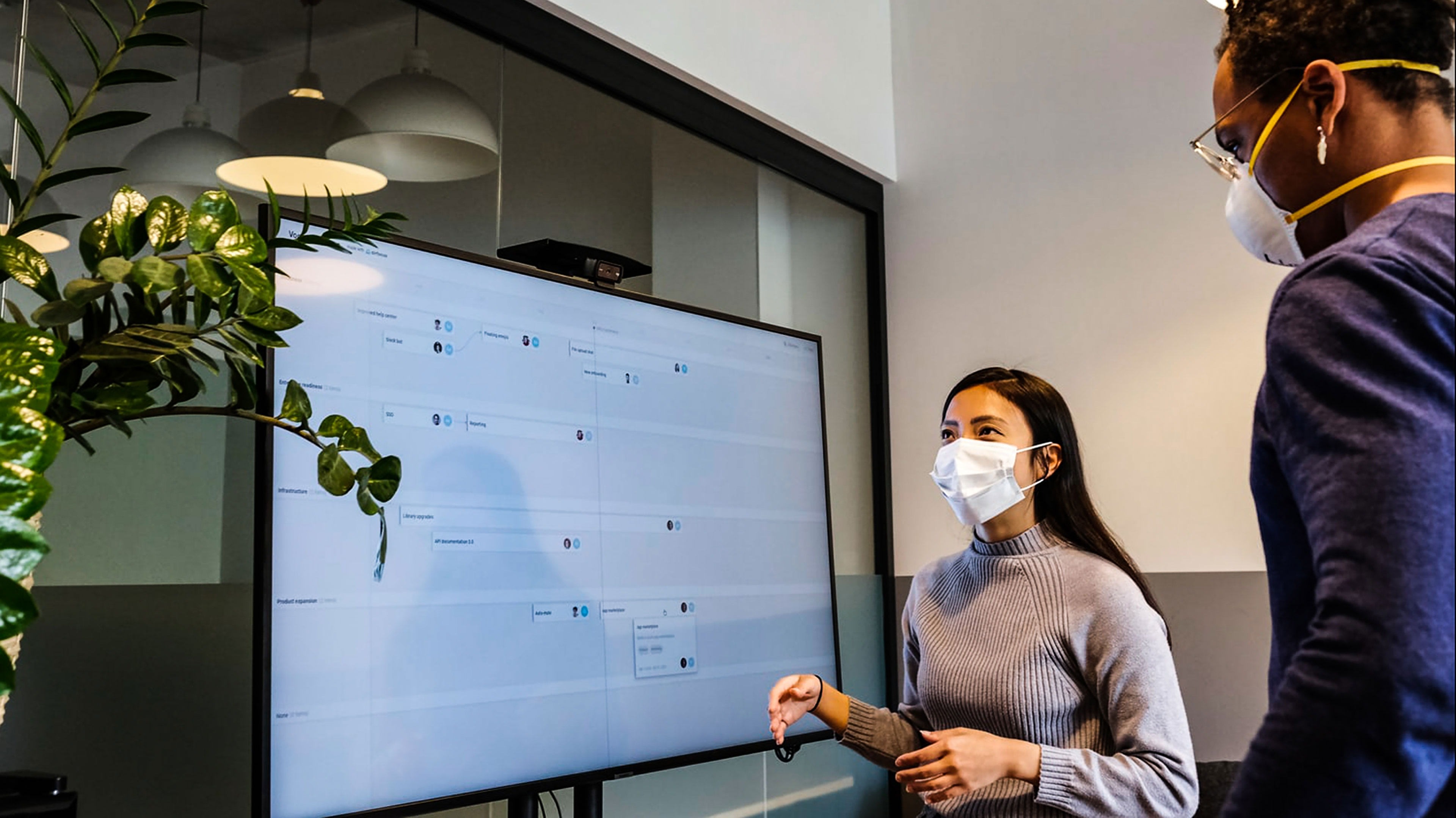 two people with masks standing in front of a large presentation screen.