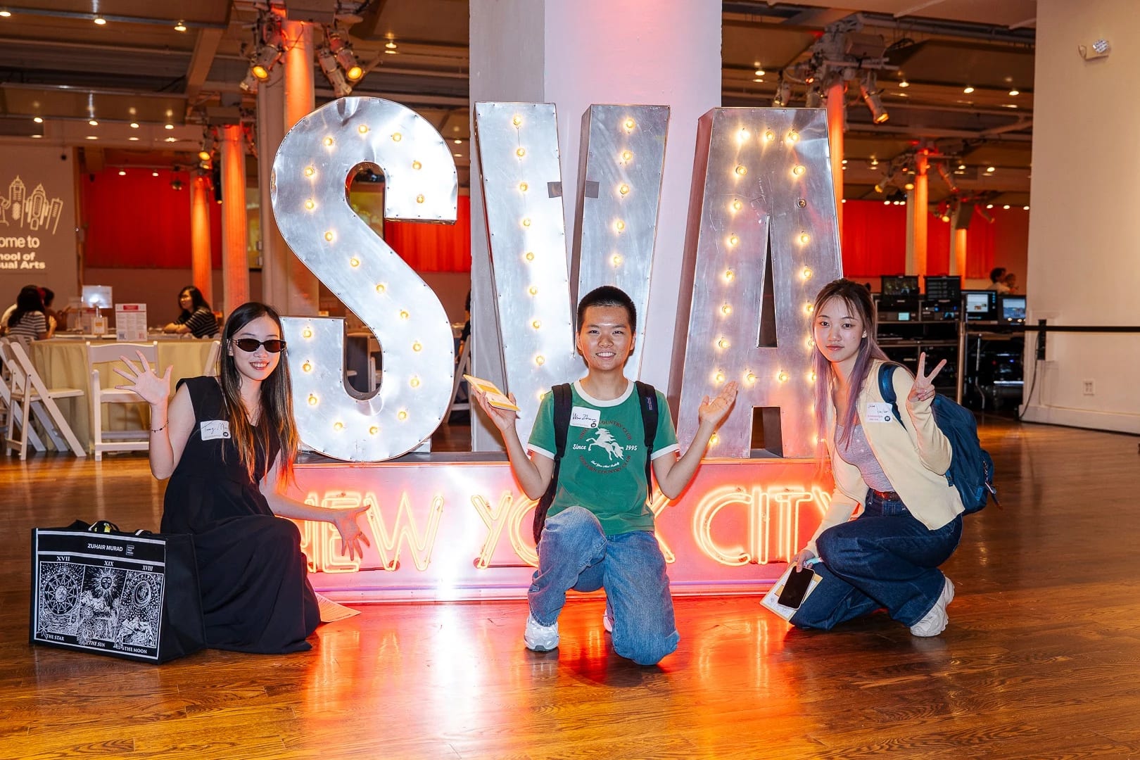 Three students pose in front of a large illuminated “SVA New York City” sign at an indoor welcome event.