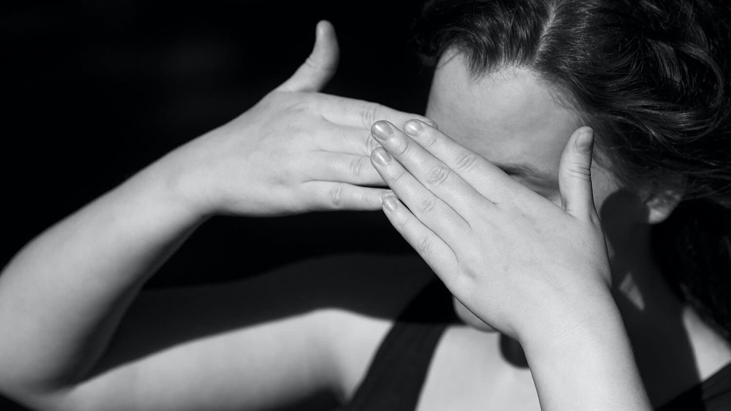 In this black-and-white photo, a person shields their eyes from the sun with their hands. 