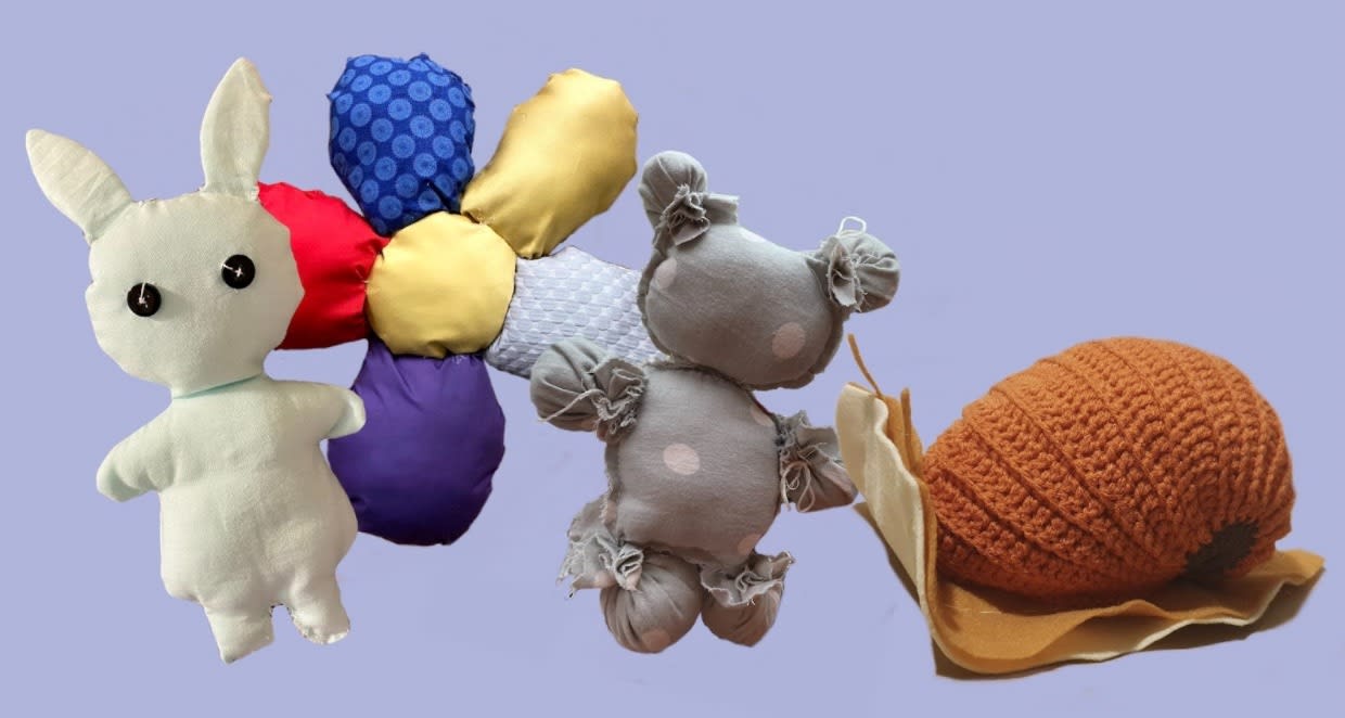 Four multi-colored, plush objects of various shapes and sizes grouped in front of a lavender backdrop.