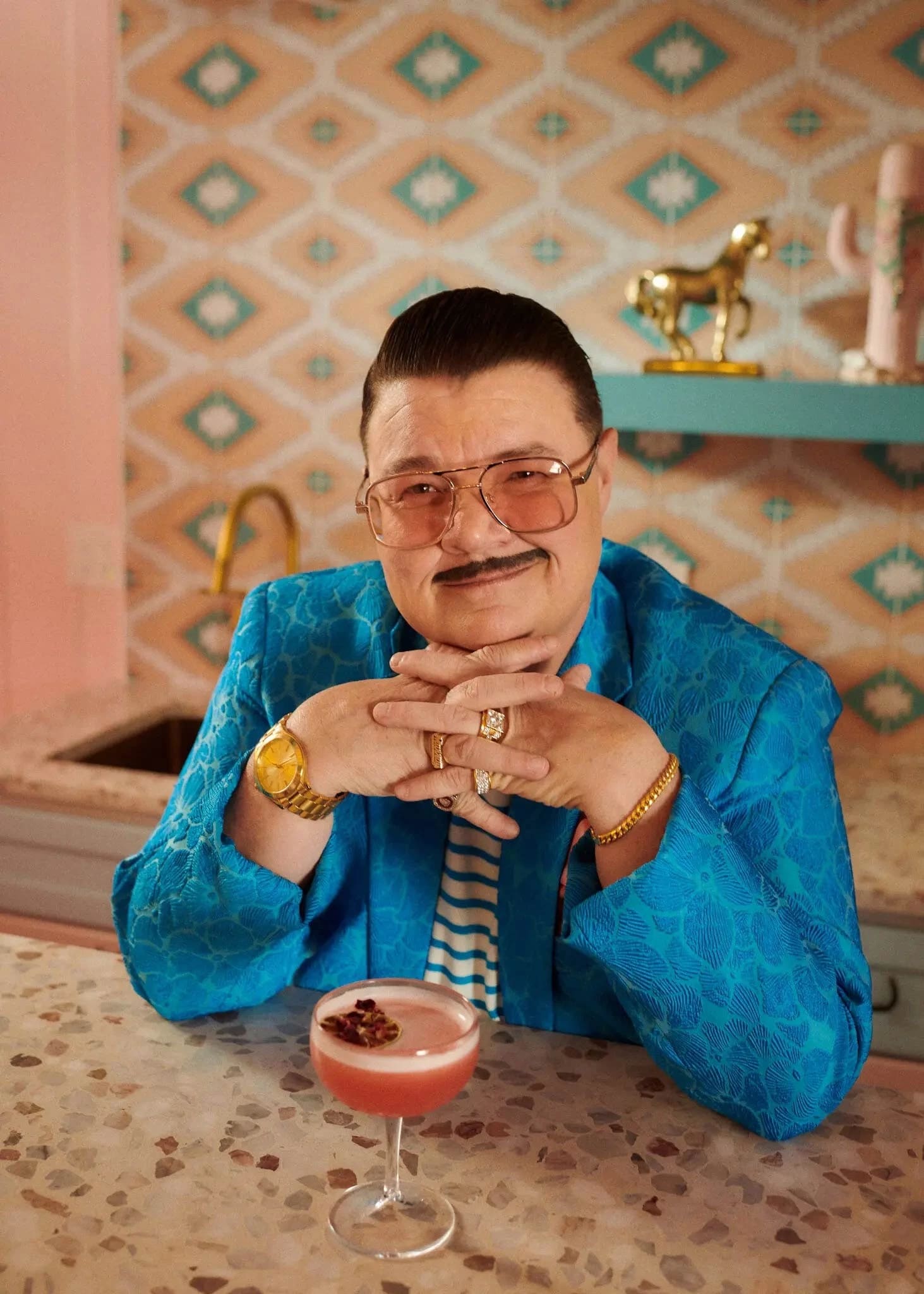 A man with pencil thin mustache and sunglasses, wearing a electric blue suit smiles at the camera.