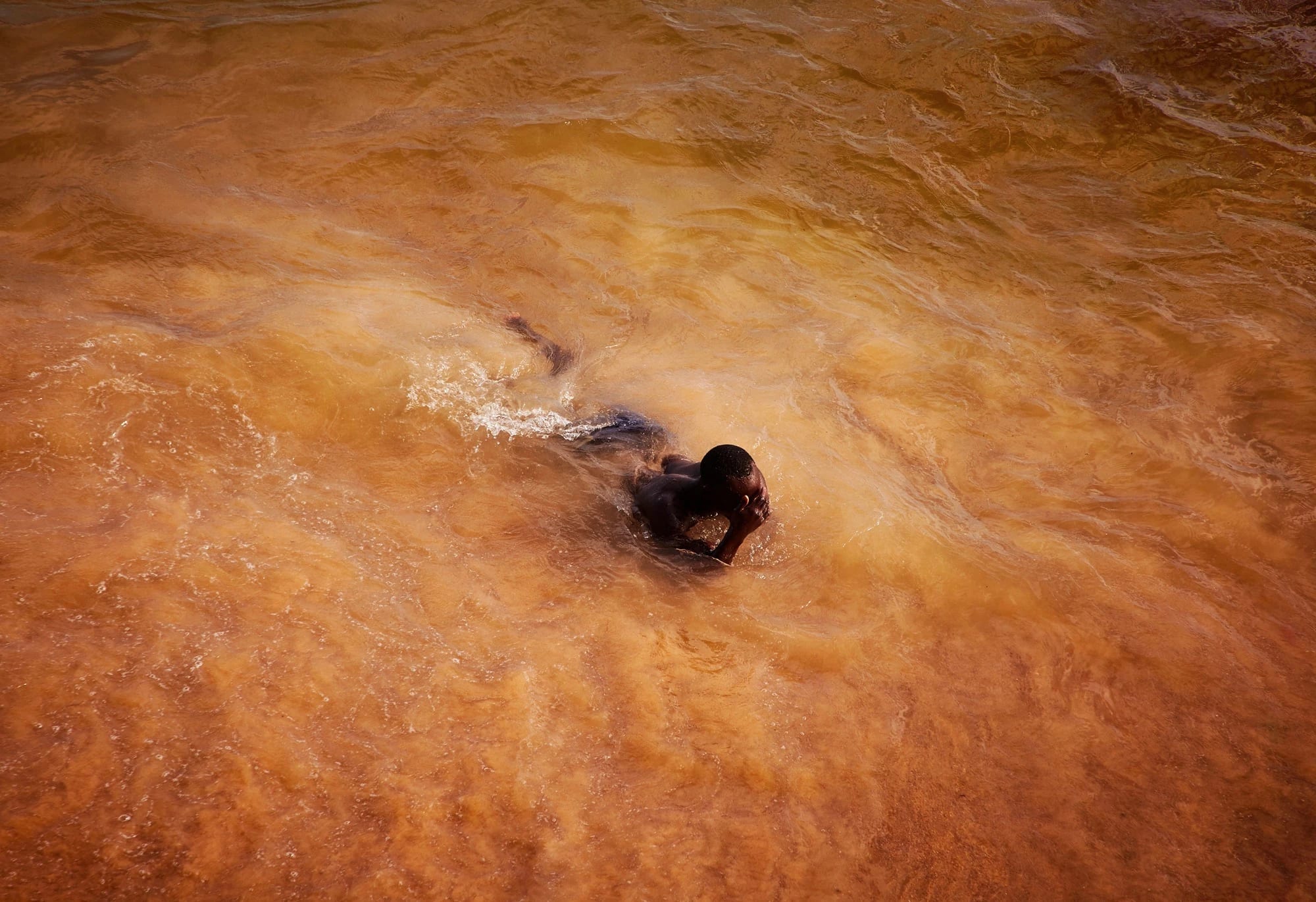 In this color photograph, a man emerges from the ocean, which has taken on a vibrant orange-ish cast in rural Saly, Senegal.