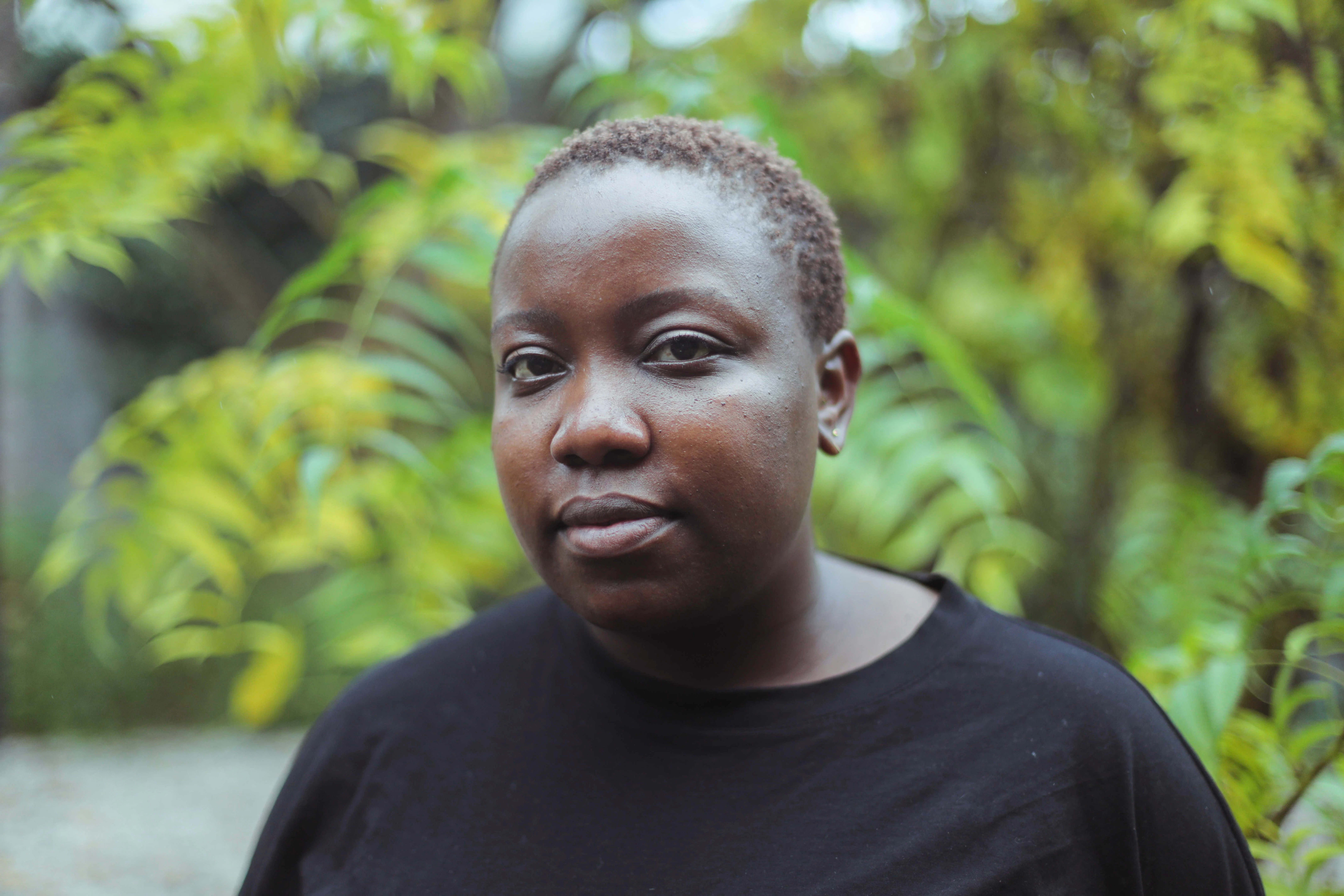 A photo of Yolande van der Heide from the shoulders up. She is wearing a black shirt and her hair is short. Behind her are big, green plants.