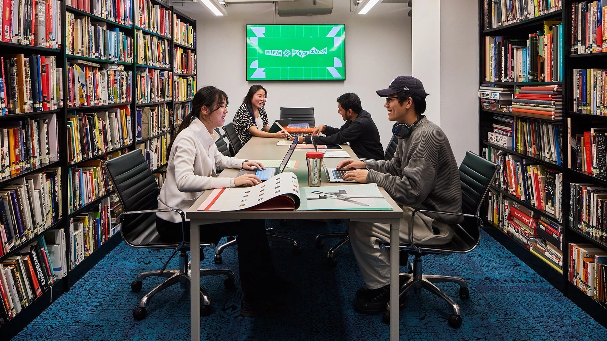 Four Students working at library table surrounded by colorful books on shelves. A green computer screen is in the background