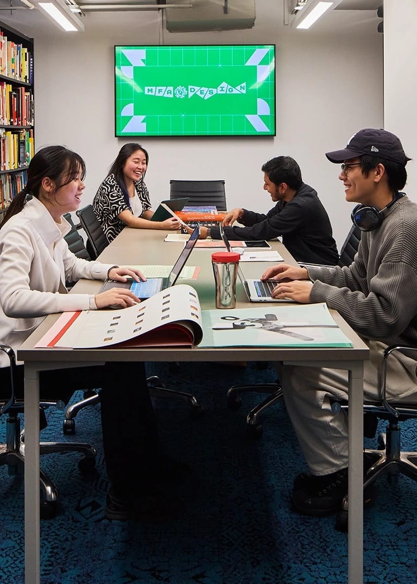 Four Students working at library table surrounded by colorful books on shelves. A green computer screen is in the background