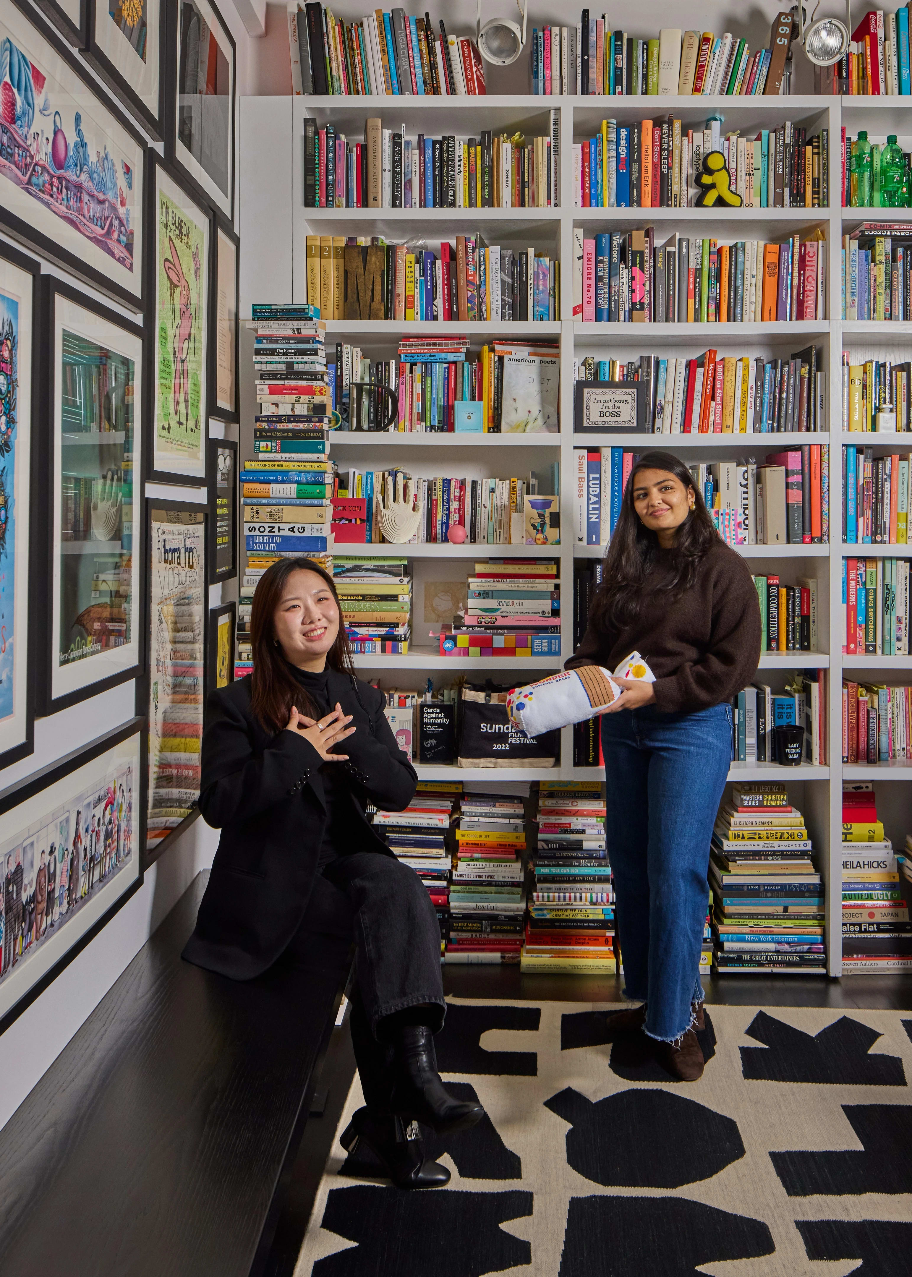 Two female students in a library room surrounded by vibrant colorful design books and art objects