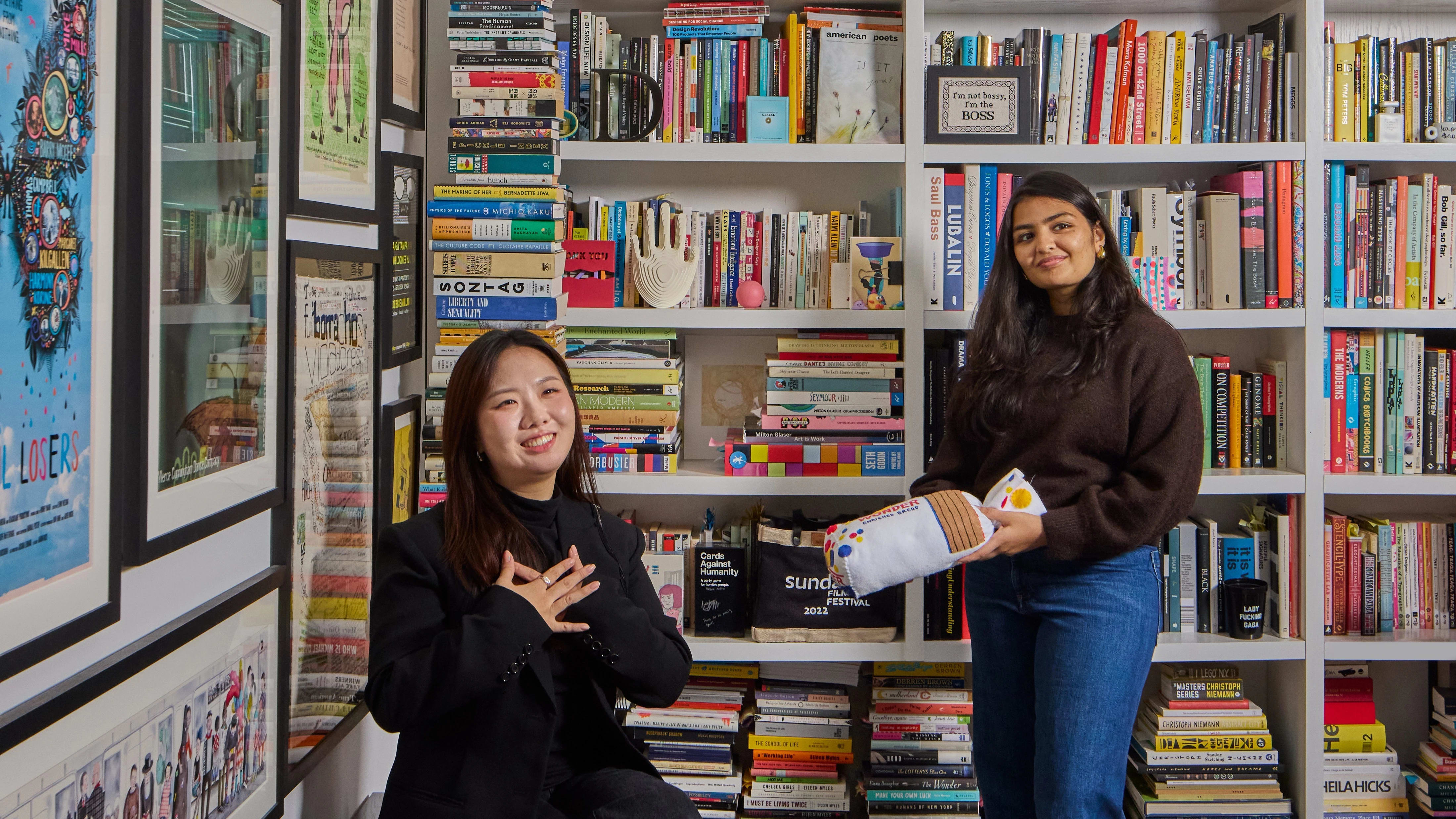 Two female students in a library room surrounded by vibrant colorful design books and art objects