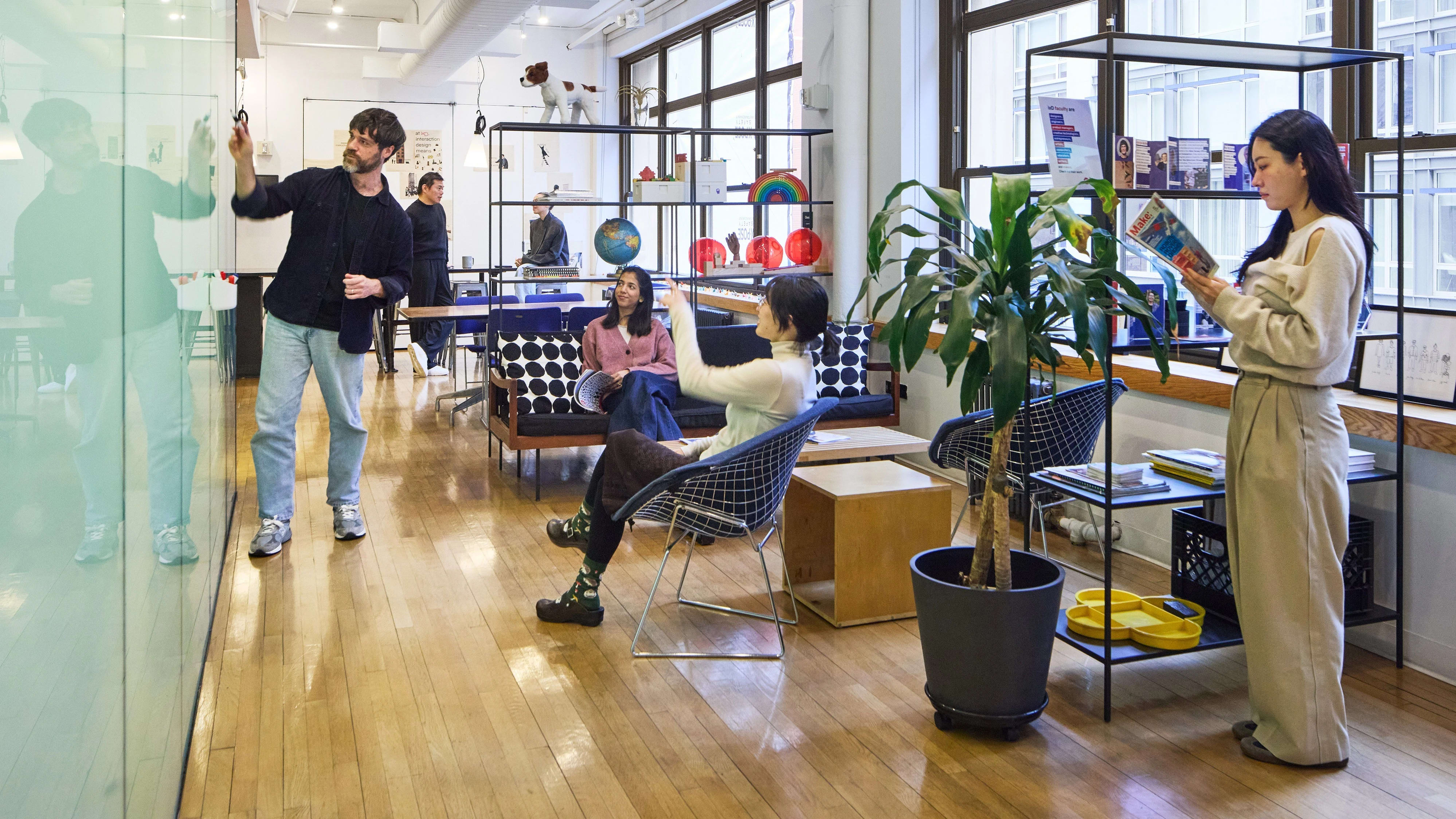 Six students in various locations of a large bright room with window, lounge furniture art objects on display. One student writes notes on a glass presentation panel