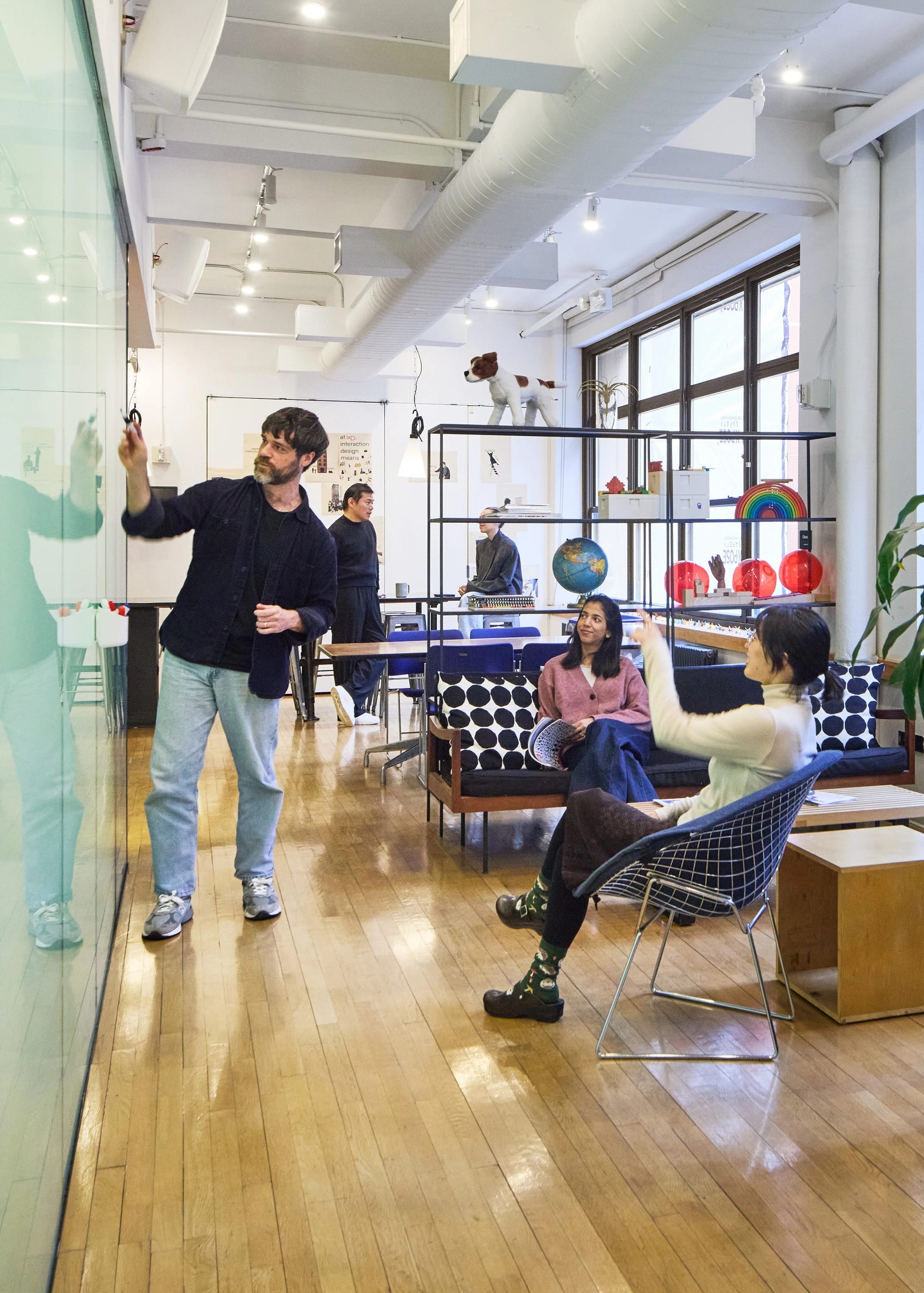 Six students in various locations of a large bright room with window, lounge furniture art objects on display. One student writes notes on a glass presentation panel