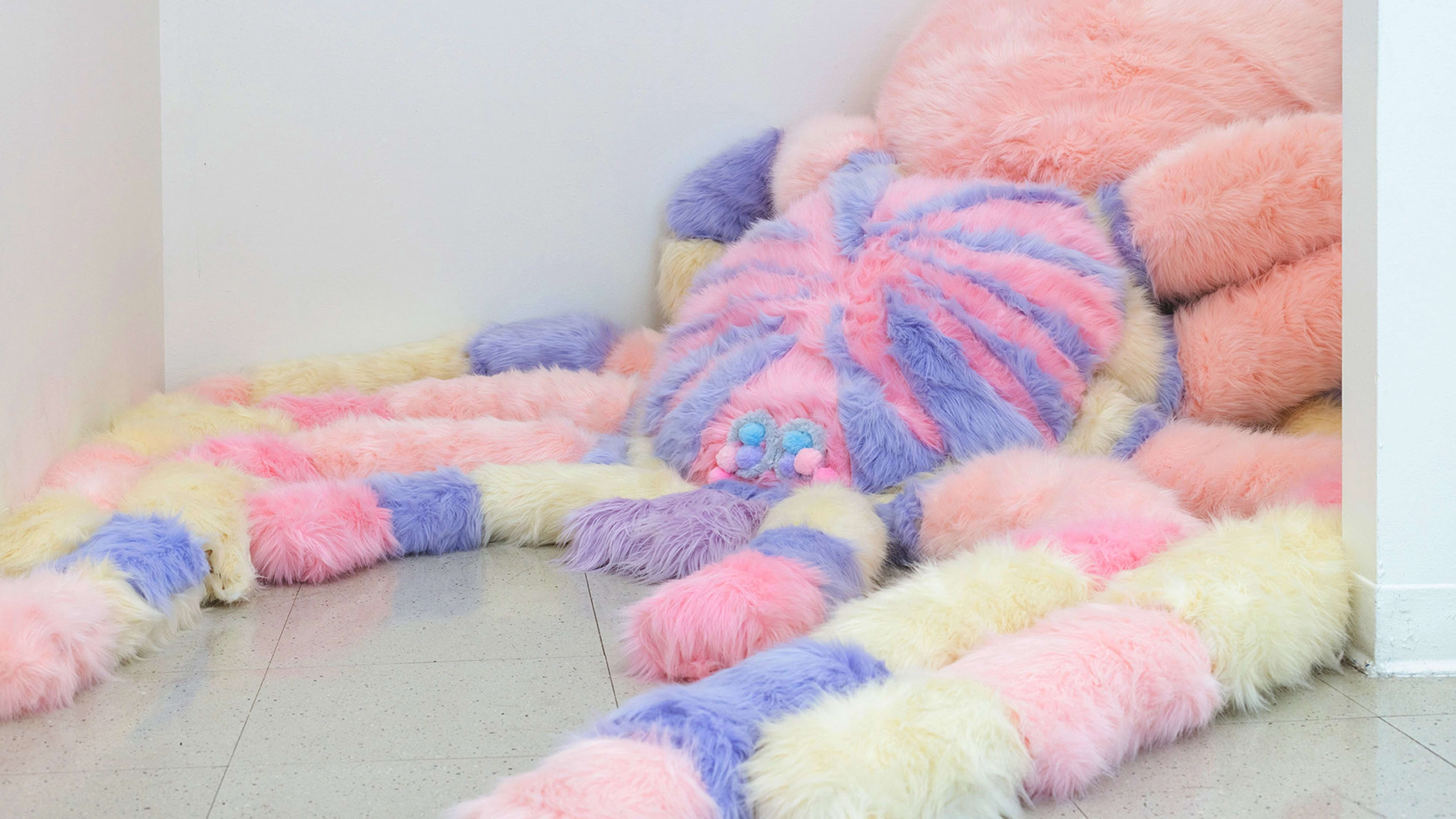 Large pink, purple and yellow soft sculpture in the shape of a fluffy spider on pale gray floor in the corner of a white room