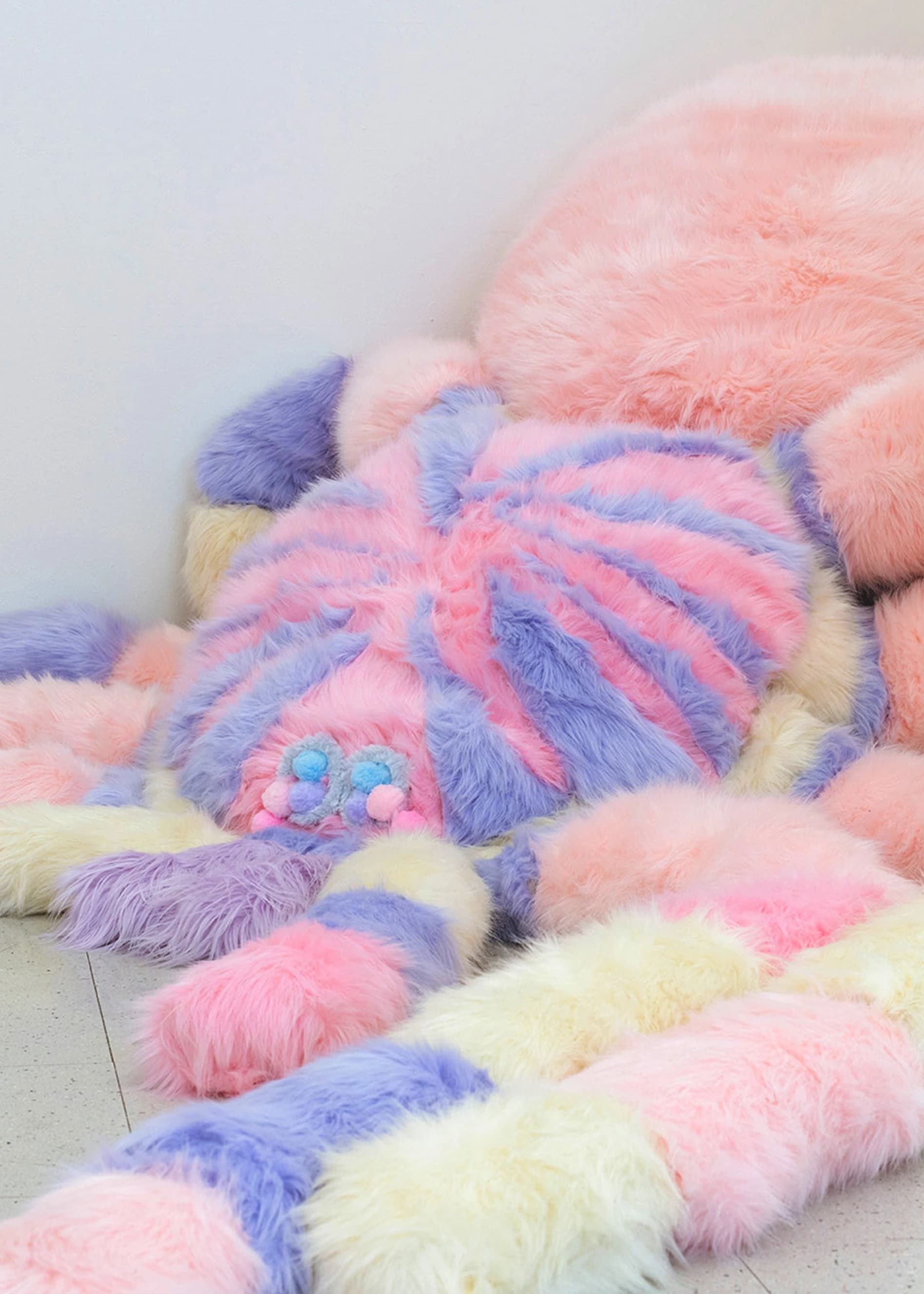 Large pink, purple and yellow soft sculpture in the shape of a fluffy spider on pale gray floor in the corner of a white room