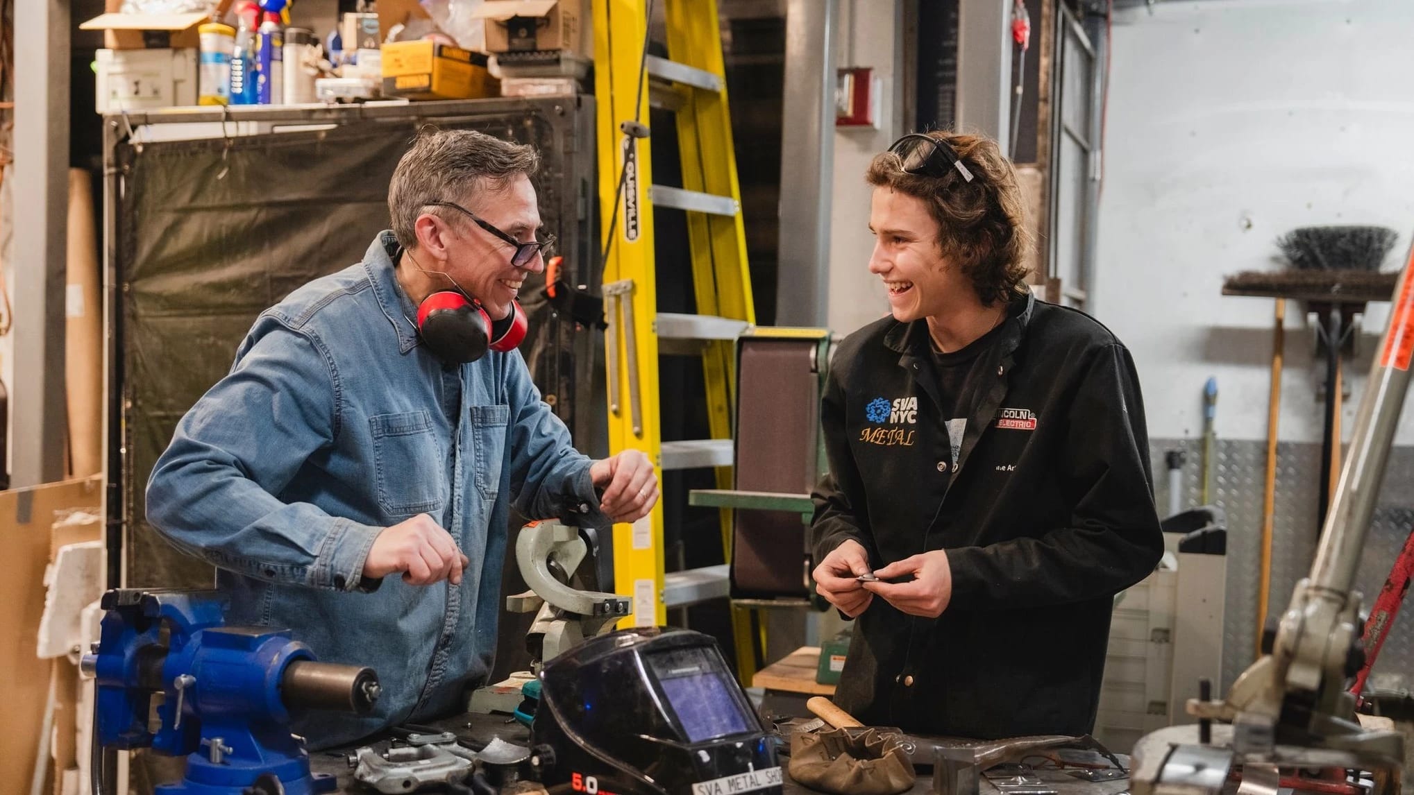 Male student working beside an instructor in metal fabrication shop with tools and equipment on their desk