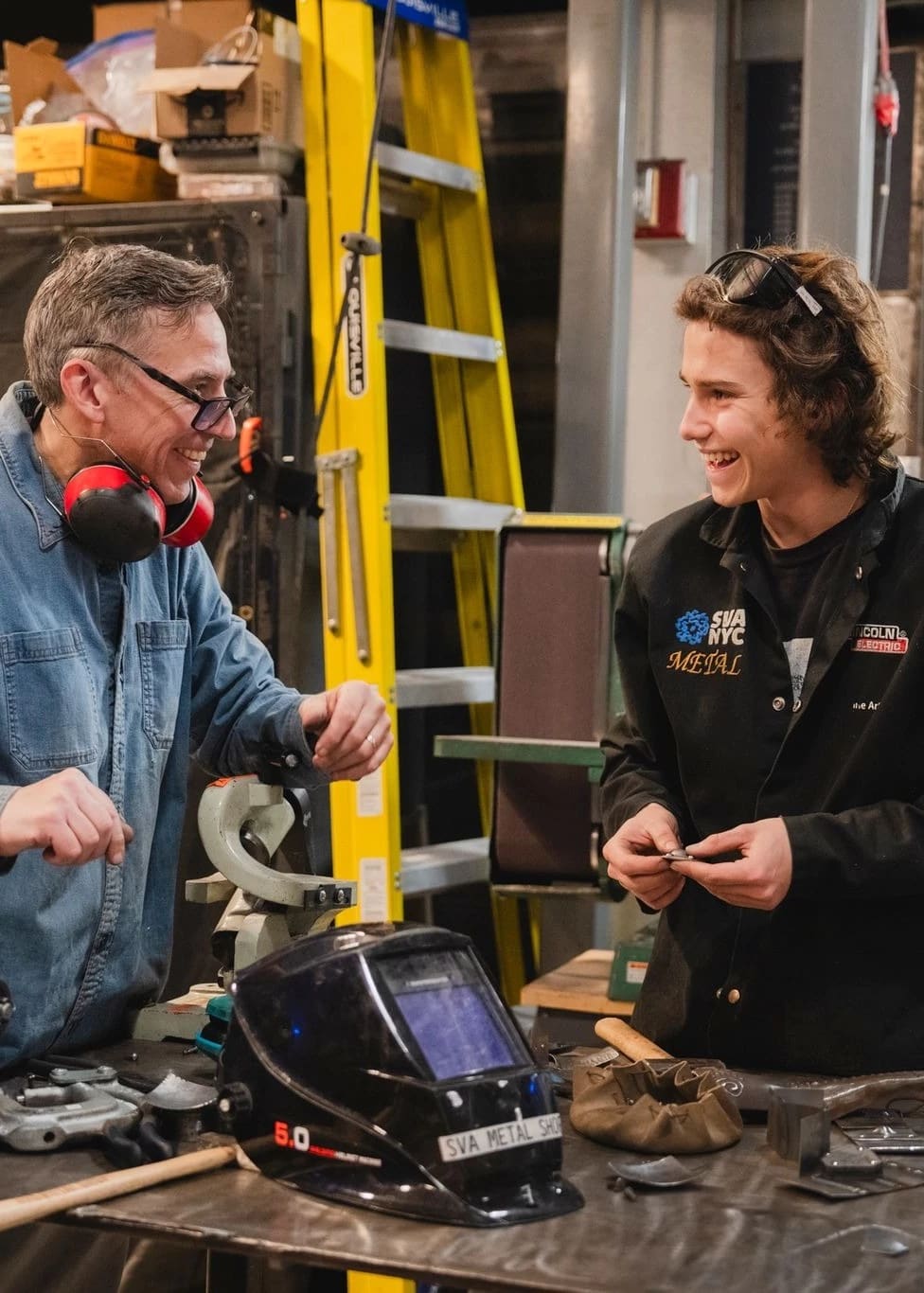 Male student working beside an instructor in metal fabrication shop with tools and equipment on their desk