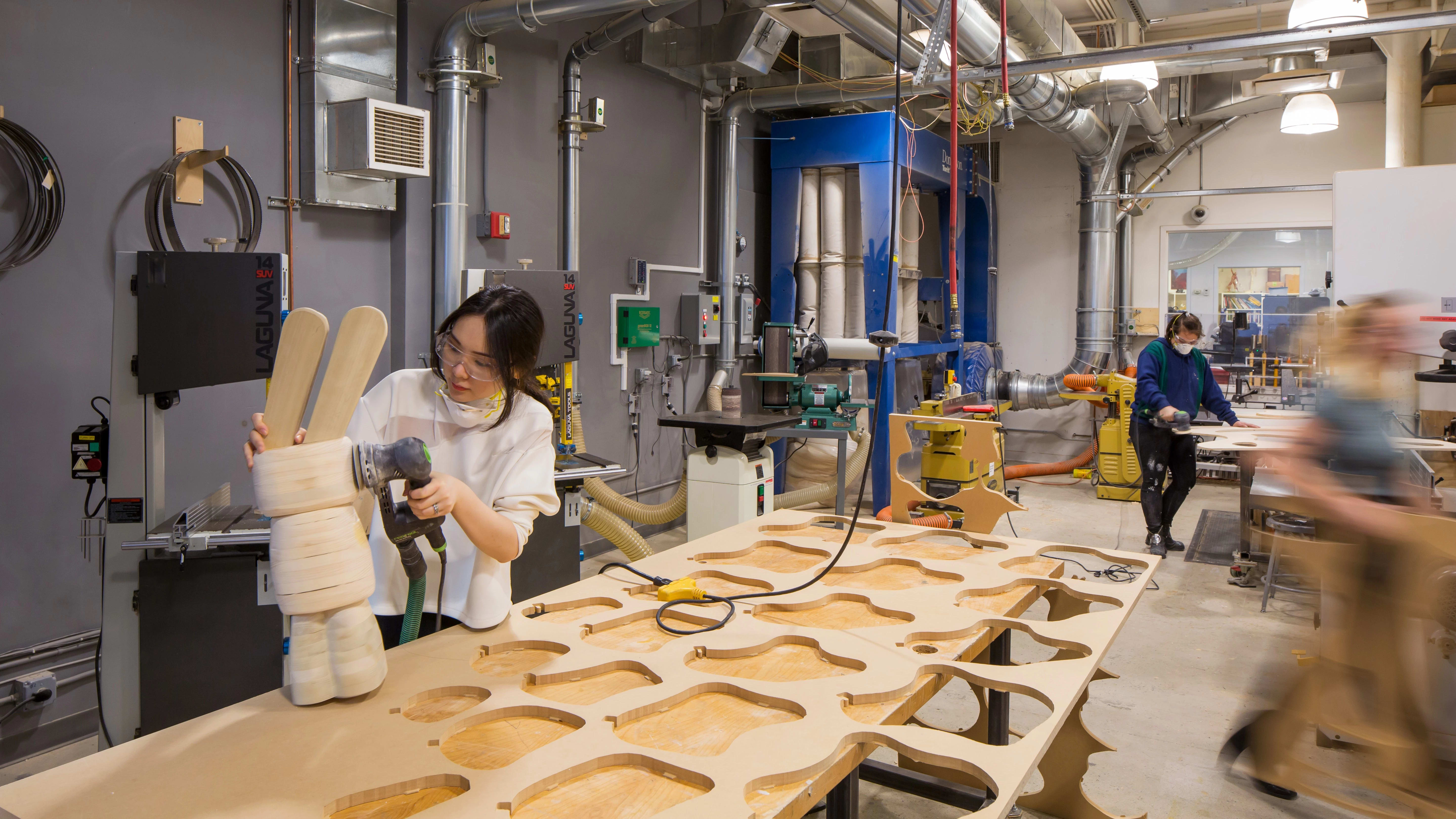 Female student in wood working studio sanding a layered wood sculpture in the shape of a rabbit many cut out wood shapes beside her