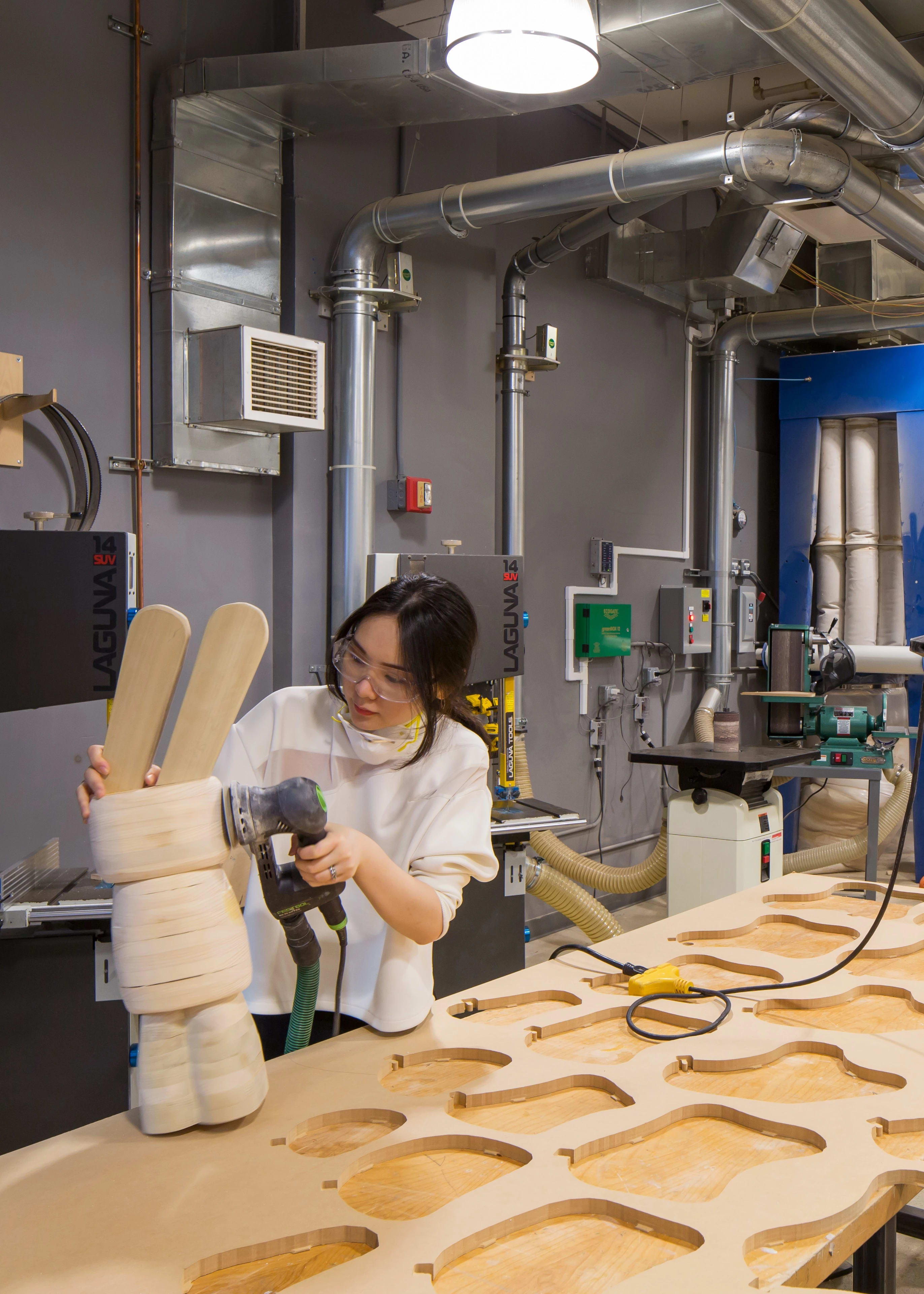 Female student in wood working studio sanding a layered wood sculpture in the shape of a rabbit many cut out wood shapes beside her