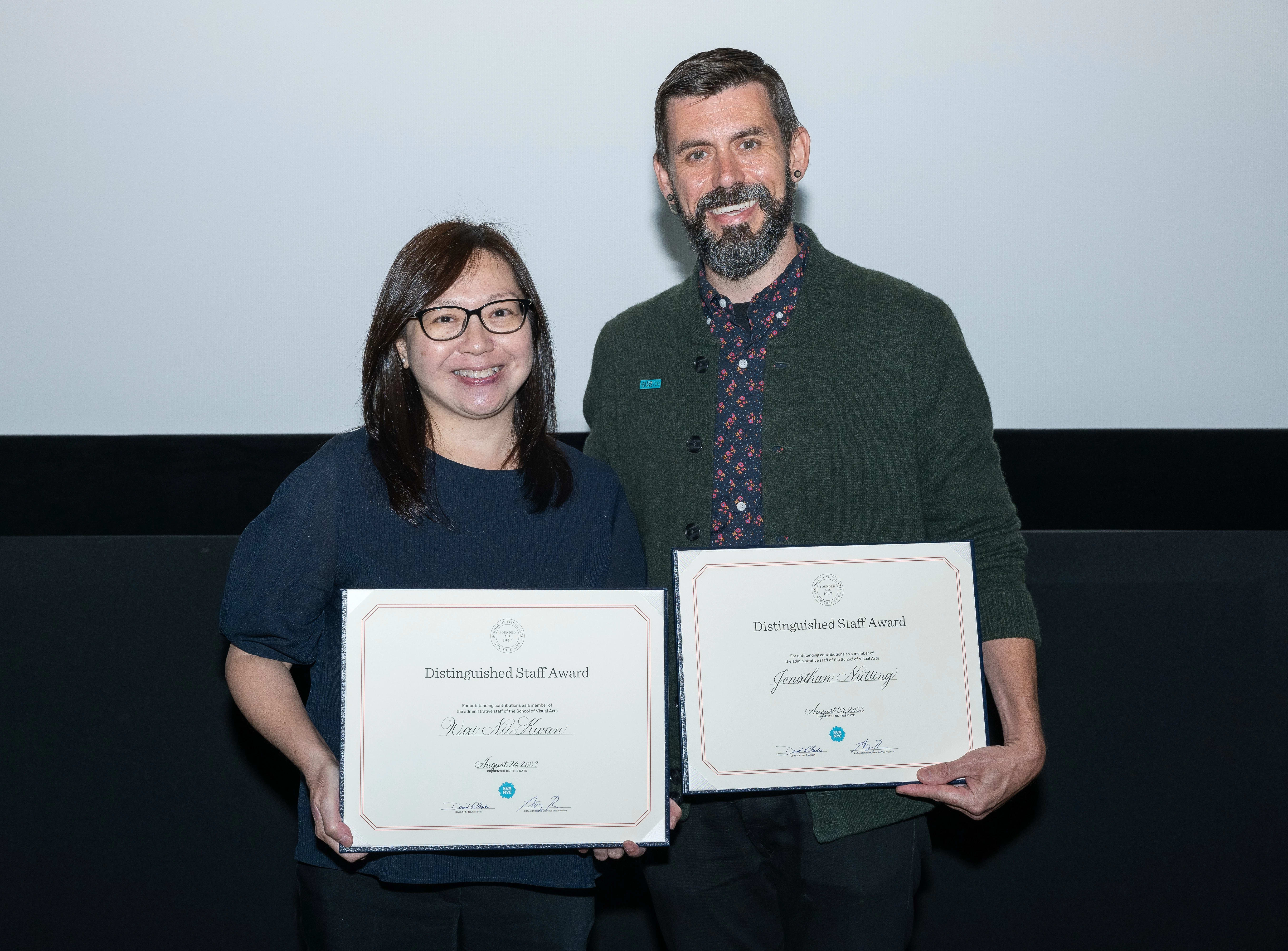 Two people stand on a stage holding awards, they are smiling and posing for the camera