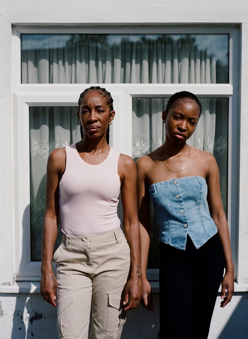 A photo of two women standing in sunlight in front of a large window with white curtains inside