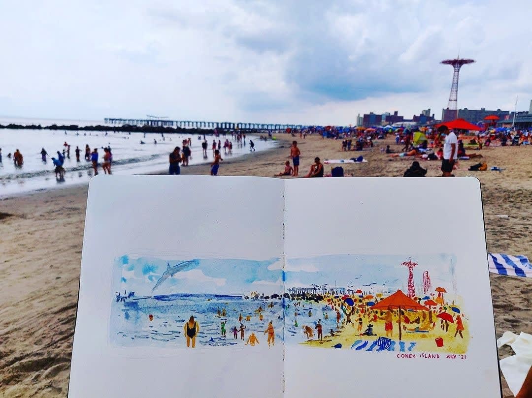 Photo of the beach at Coney Island, with the water to the left. People are in the water and on the beach, and the amusement park is in the distance. In the foreground, a sketchbook is open to a colorful sketch of the beach and park.