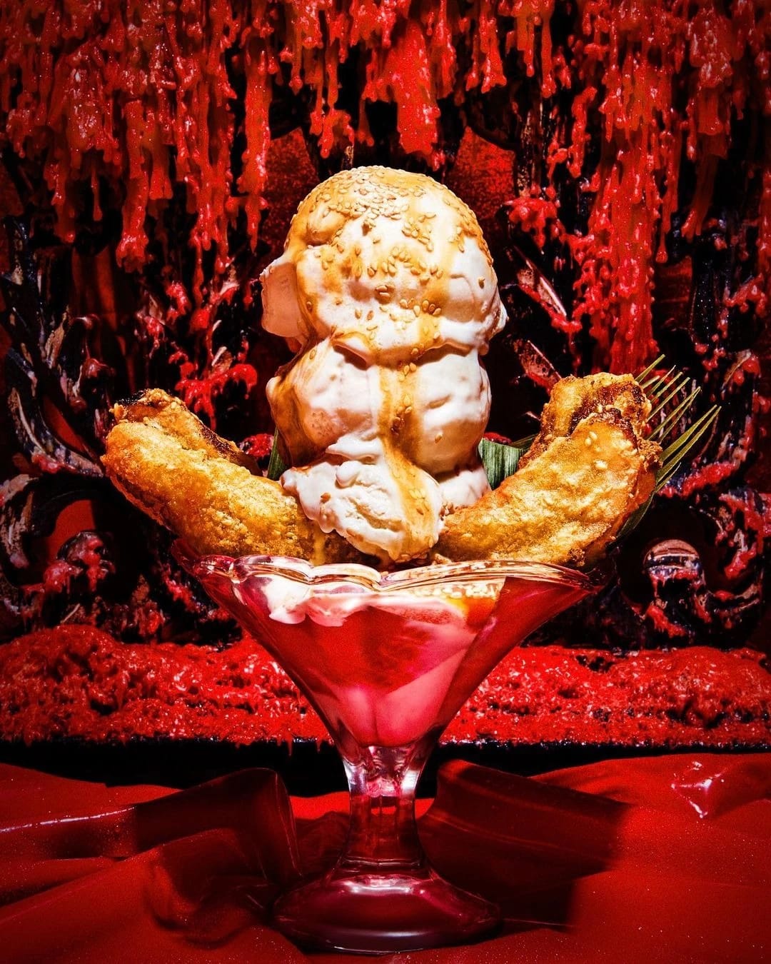 A photograph of white ice cream on top of deep fried food in a sundae glass. The background is red and looks to be covered in dripping wax. The light illuminating the food is red too.