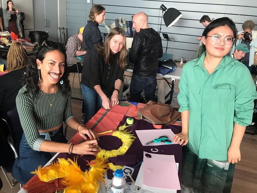 Three people sit around a table that is covered with quilting supplies including felt, scissors, feathers, and fabric