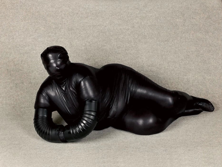 a person in a full-body black latex suit reclining in front of a gray background