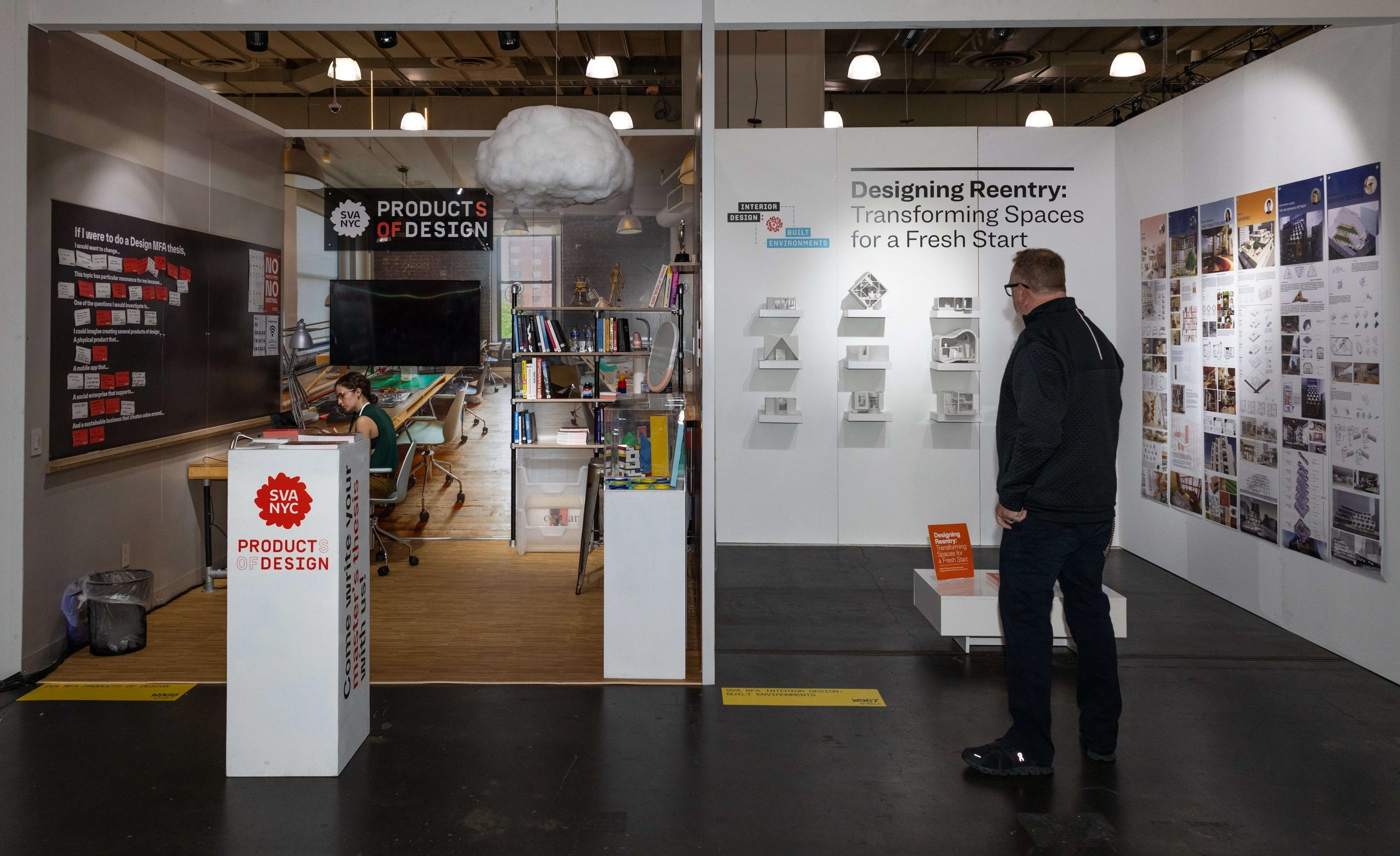 A man looks at a booth that has student projects in it next to another booth with a women sitting at a desk towards the back