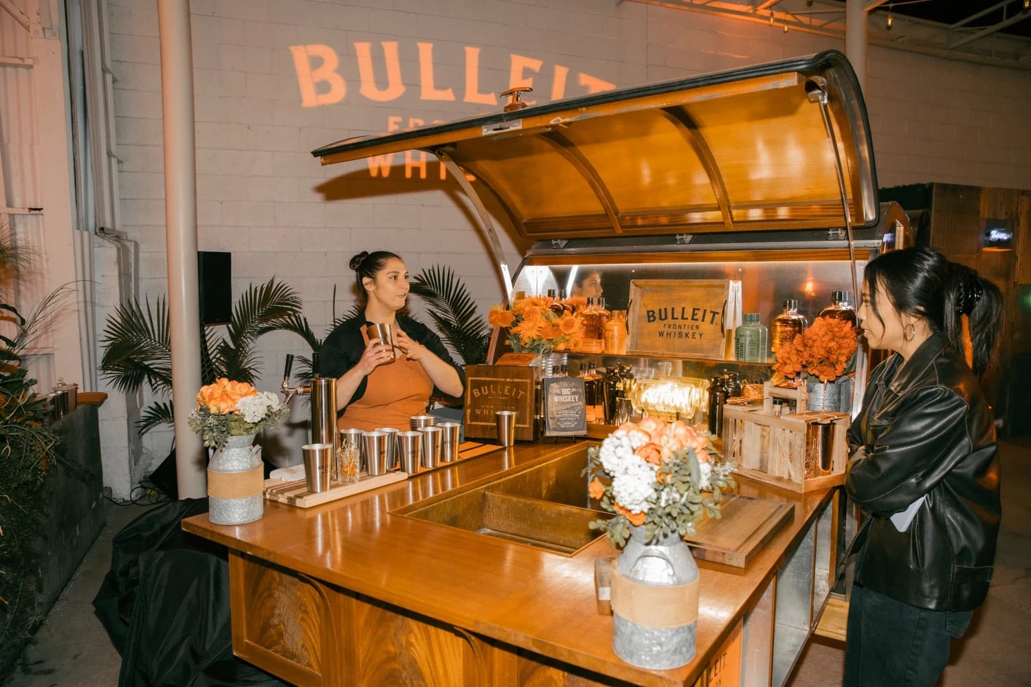 Two women stand at a bar made of wood. One woman is ordering a drink and the other is making it.