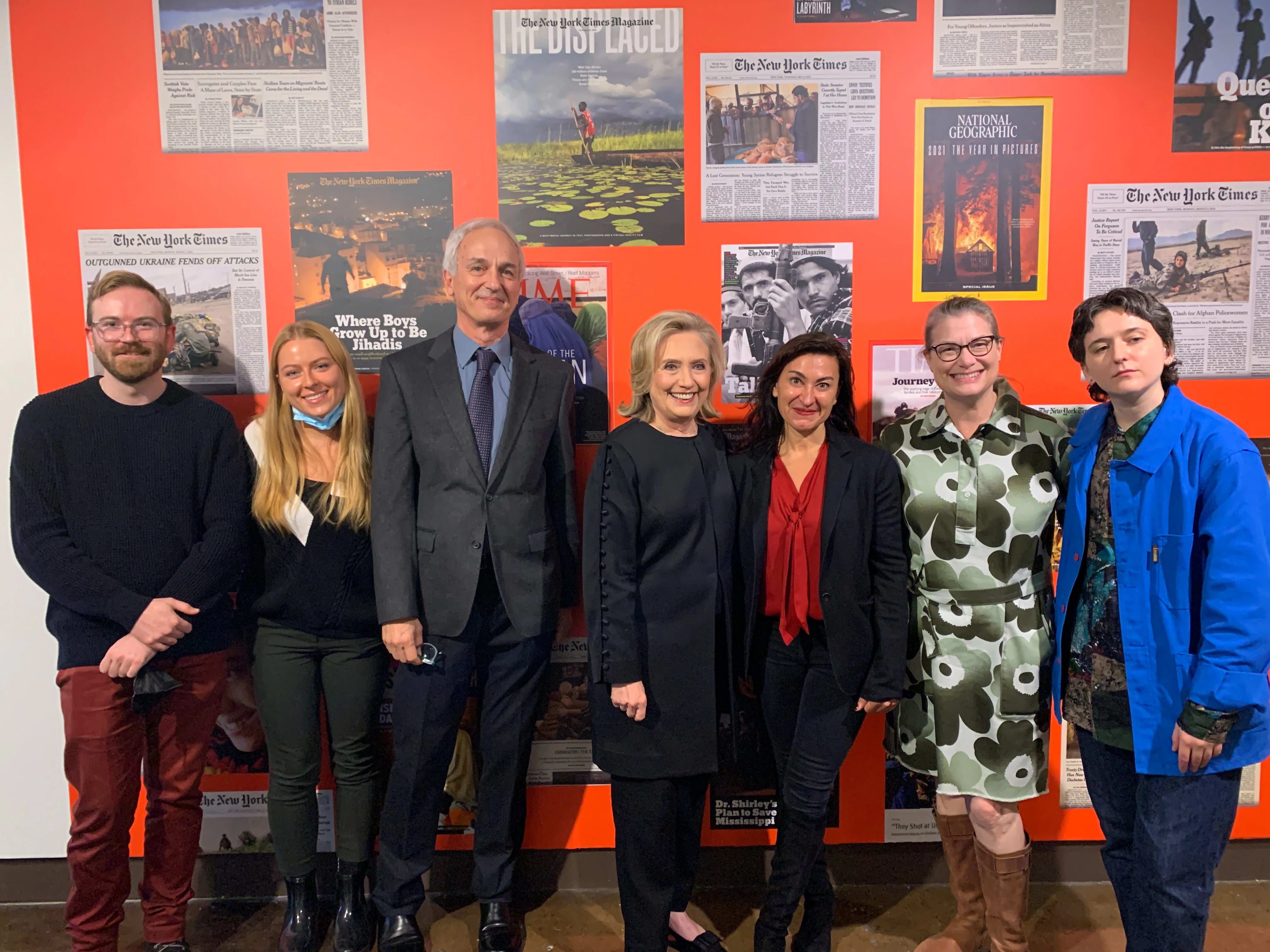 A photo of a group of seven people standing side by side in front of a red wall of photographs. In the center stands Hillary Clinton.