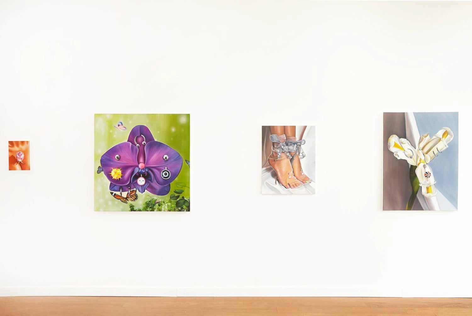 A wall of 4 paintings. Left to right: a small painting of a flower, a larger painting of a purple flower pierced with jewelry, a painting of feet, and a painting of another flower that has been pierced.  