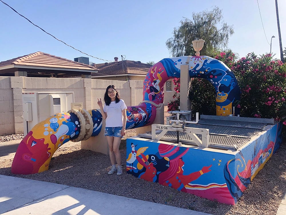 A photo of the artist, Yuke Li, next to a waterpipe painted with a mural designed by her.