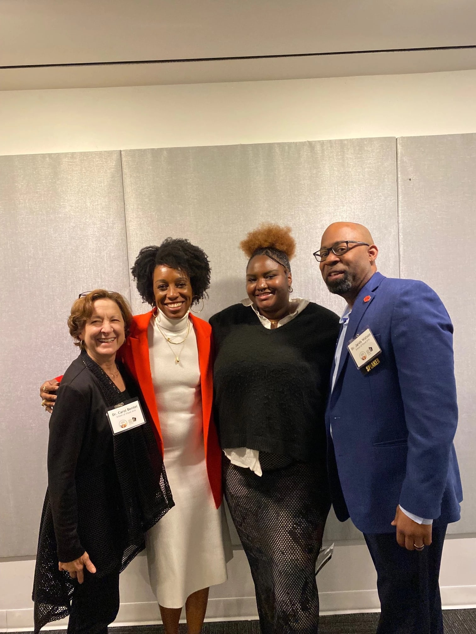 A photo of four people posing in front of a white backdrop. From left to right: a woman in a black outfit with short brown hair, a woman in a white dress and red cardigan with curly black hair, a woman in a black top and pants with a necklace on and her hair in a bun, and a man in a blue suit with glasses.