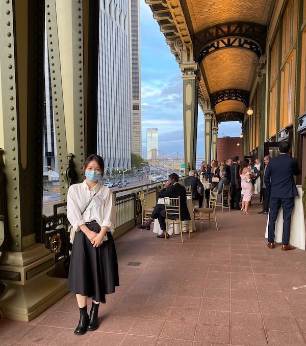 A person poses on a long outdoor balcony with high arched ceilings. A city and blue sky are seen over the balcony.