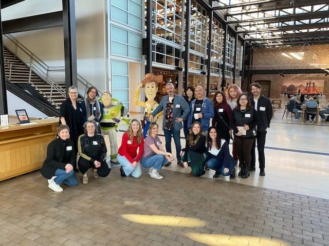 A group of people stand in a building with a tall ceiling and pose for a photo next to large figures of Woody and Buzz Lightyear from Toy Story