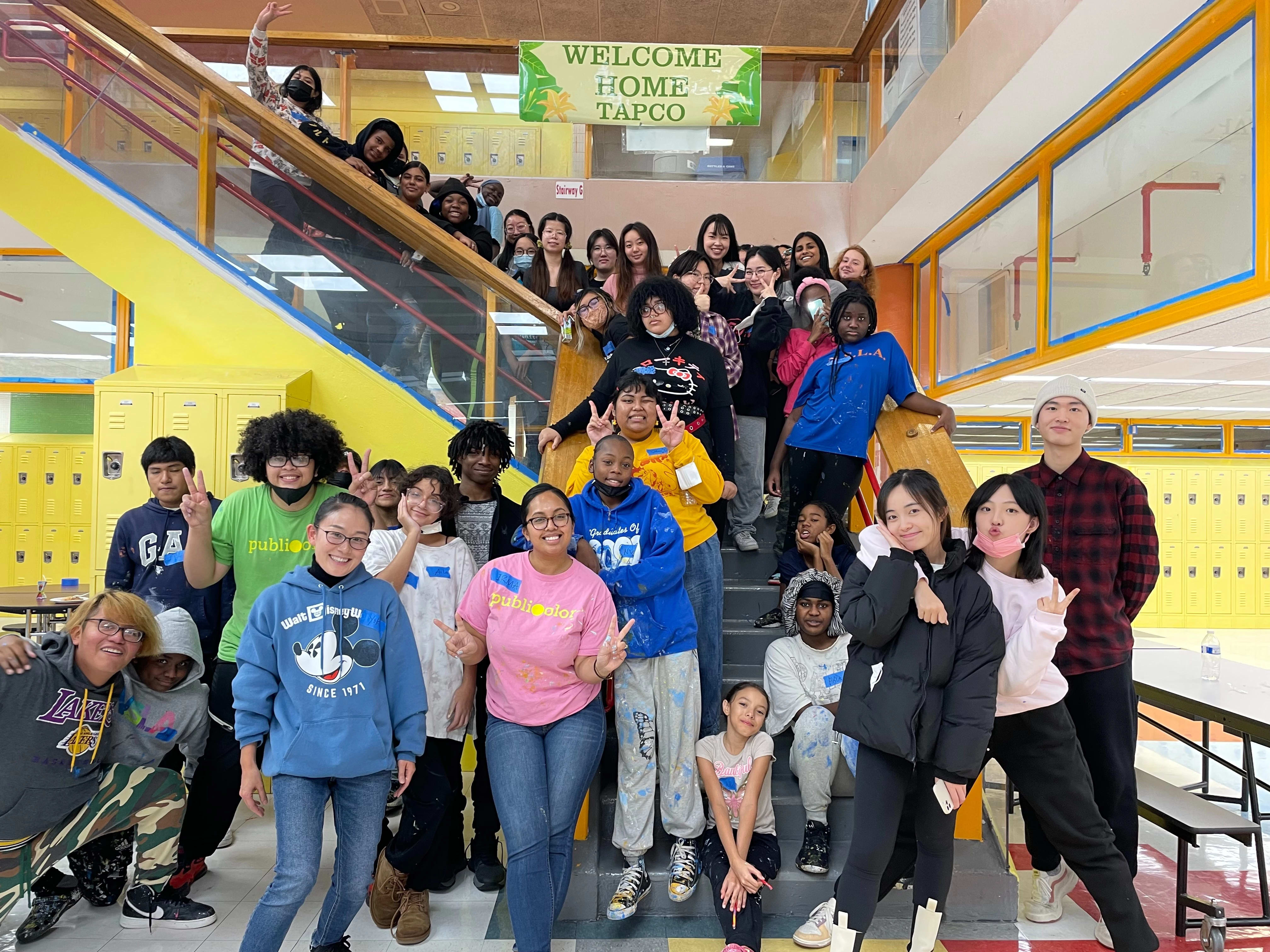 A photo of a group of young kids, filling up a staircase in the middle of a school hallway. They are surrounding by yellow walls and lockers