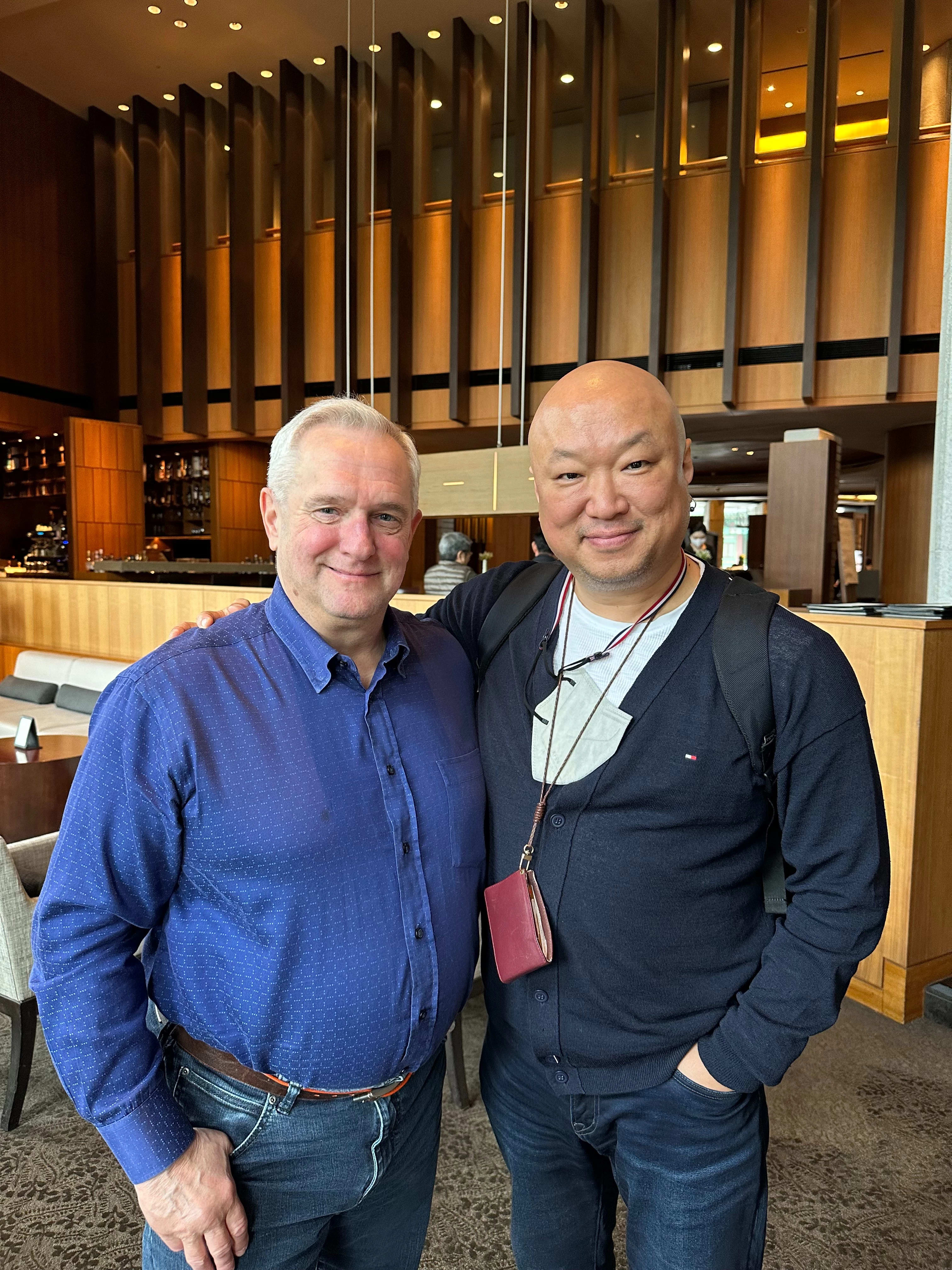 Two men stand in a hotel lobby. One is older with white hair and wears a blue button up. The other is bald and wears a dark blue cardigan.