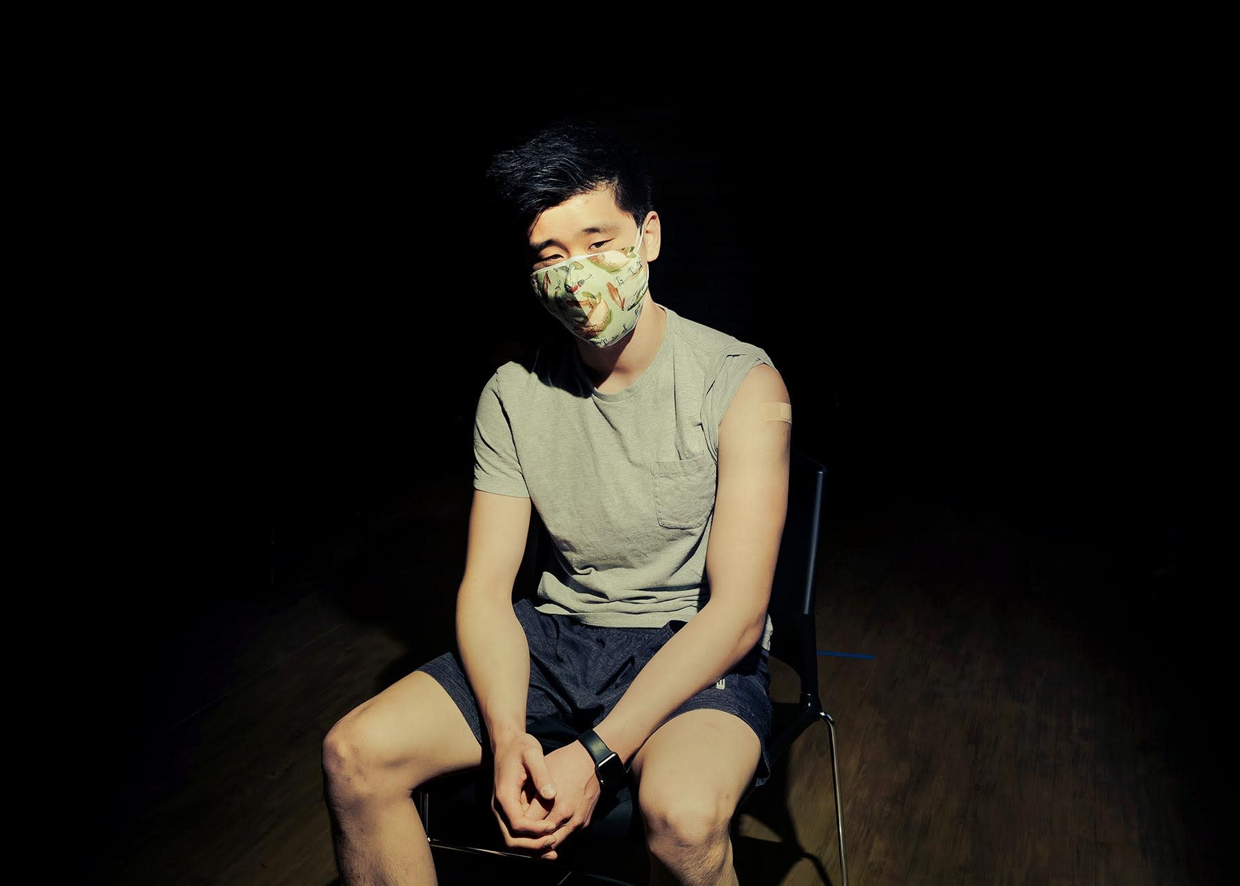 Photo of a man wearing a face mask and sitting on a folding chair. His sleeve is rolled up to his shoulder and there's a bandage on his upper arm where he received a COVID-19 vaccine.