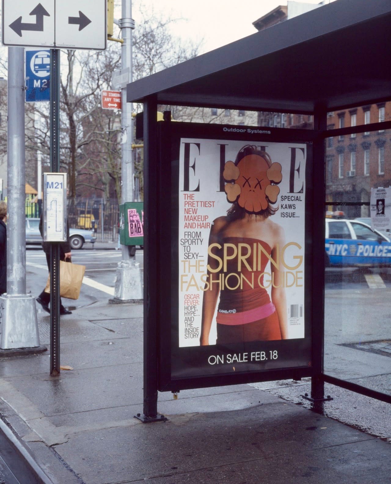 Photo of an ad in a bus stop. It is an Elle magazine cover about spring and there is a woman on the cover. The woman's face is covered by a KAWS skull and crossbones illustration