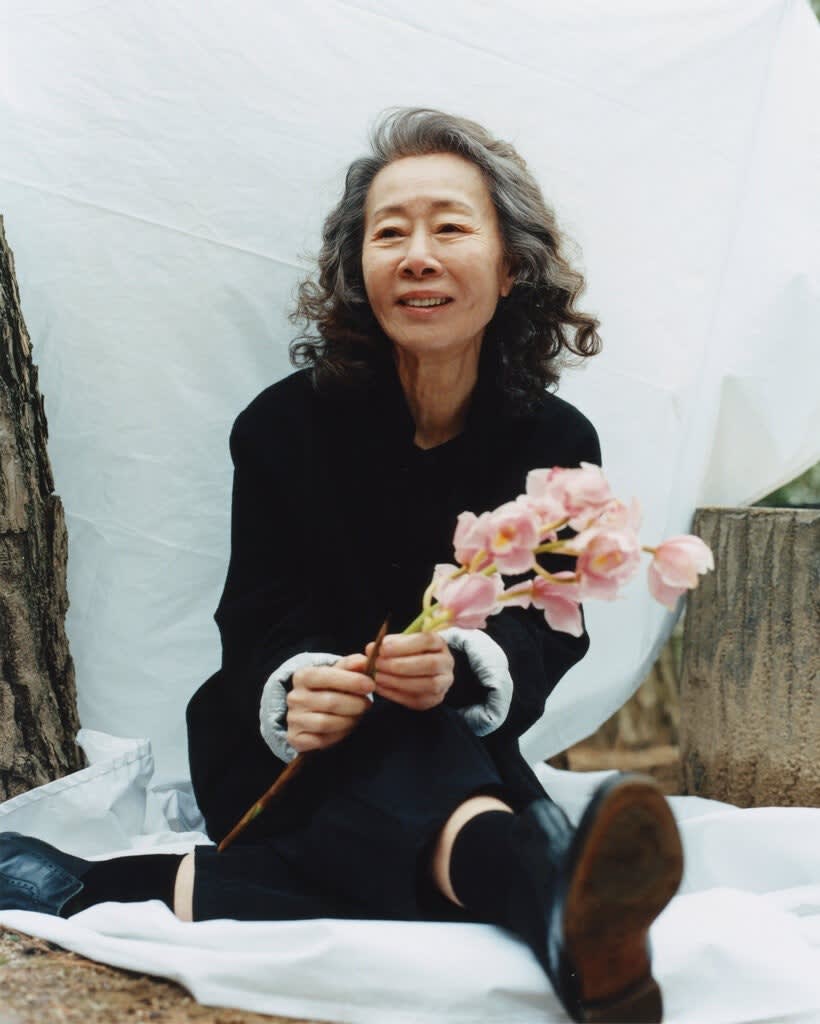 Photo of Korean actress Yuh-Jung Youn sitting on a white sheet on the ground holding pink flowers and smiling into the distance.