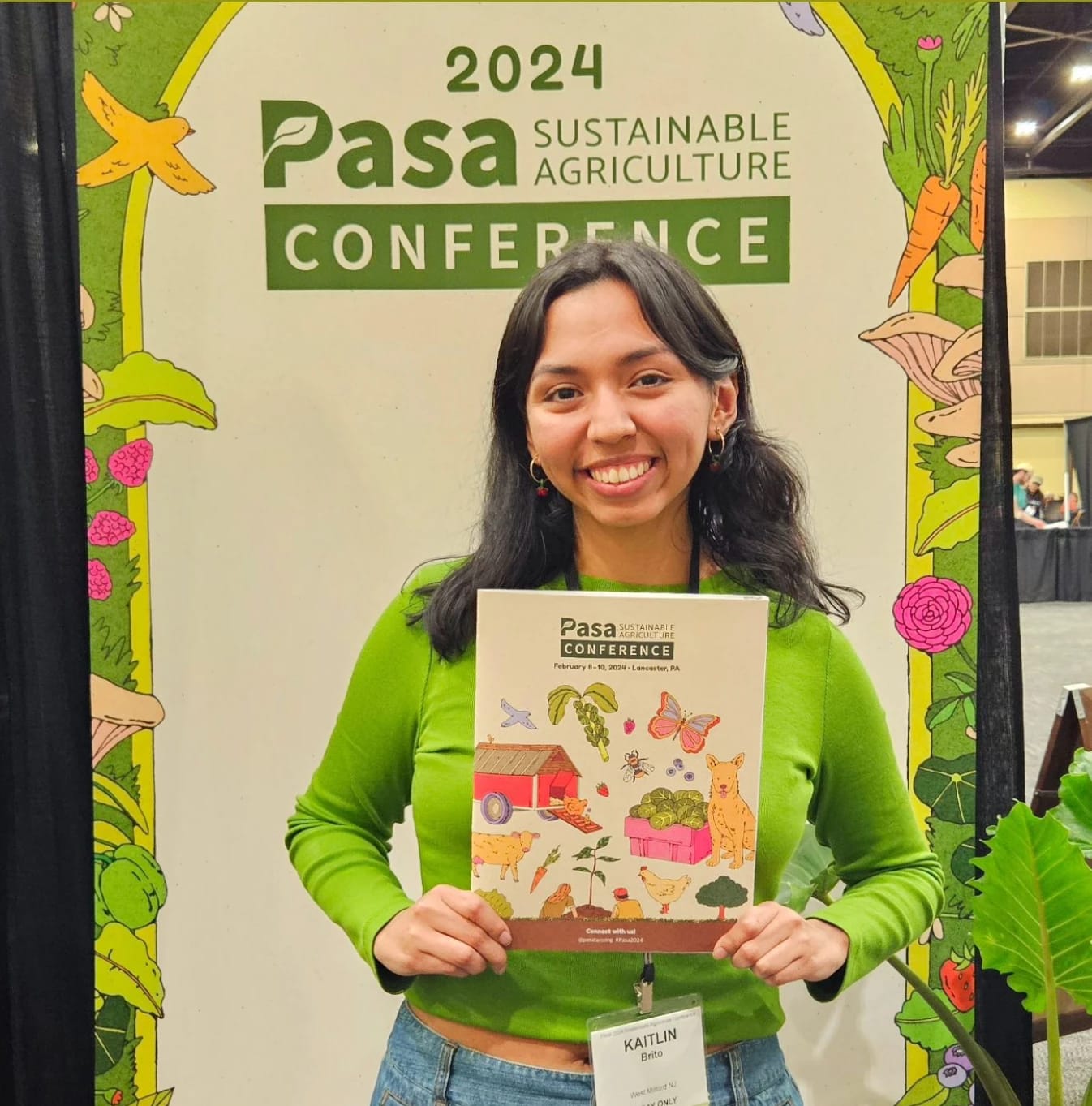 A girl in a green sweater holds a booklet with colorful illustrations on it in front of a large banner backdrop with similar illustrations 