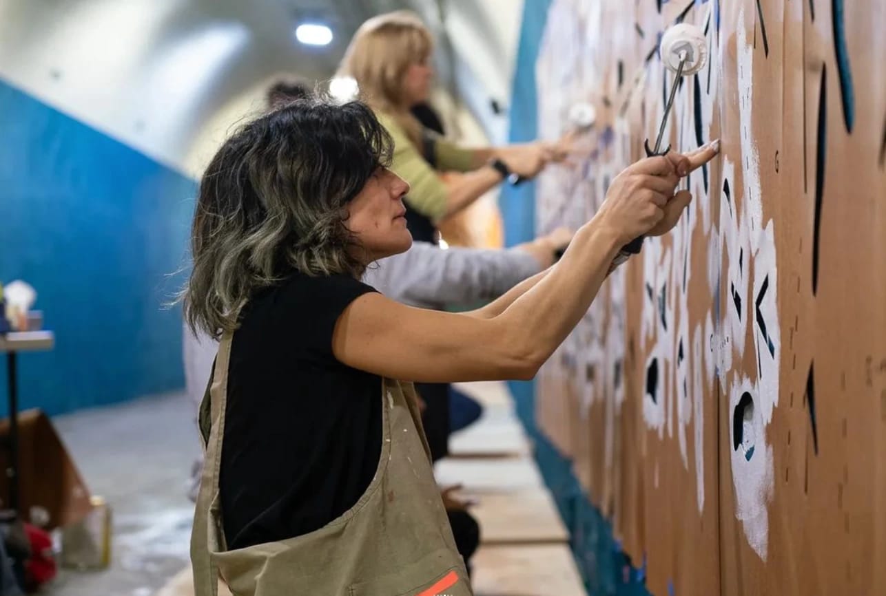 A woman paints a wall with a group of other people. There is a stencil with cut out footprints taped to the wall