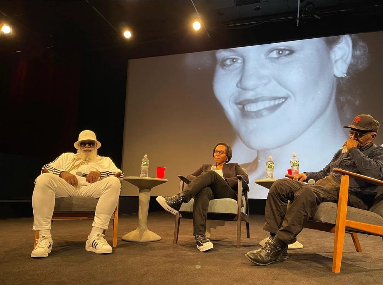 Three people sit in chairs on stage in front of a projection of a woman's face