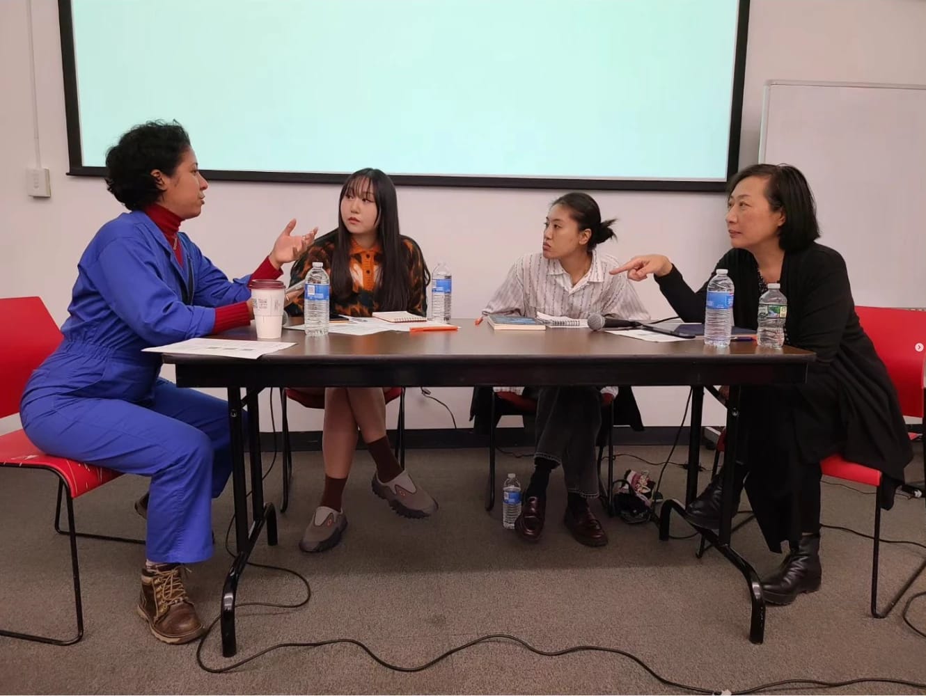 Four women sit at a folding table in front of a projector screen, they are all having a discussion together