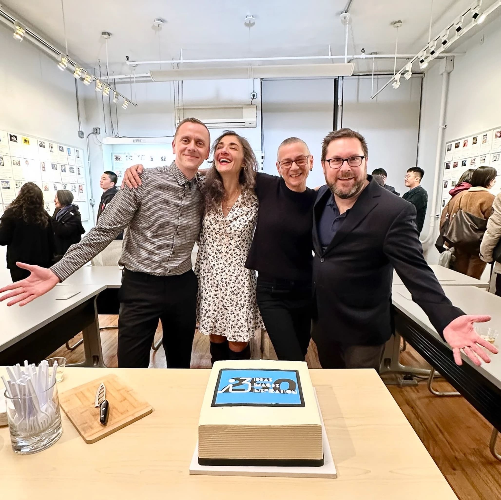 A group of four people embrace in a classroom looking happy, there is a white cake in front of them with a black and blue logo on it