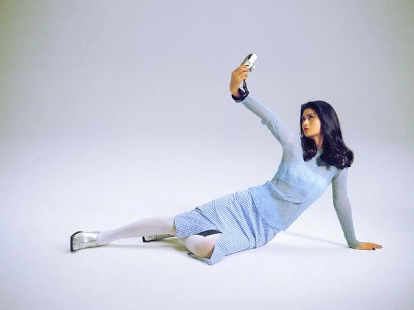 a woman in a white room laying on the ground propped up by her hand. She is wearing a blue sheer dress and taking a selfie 