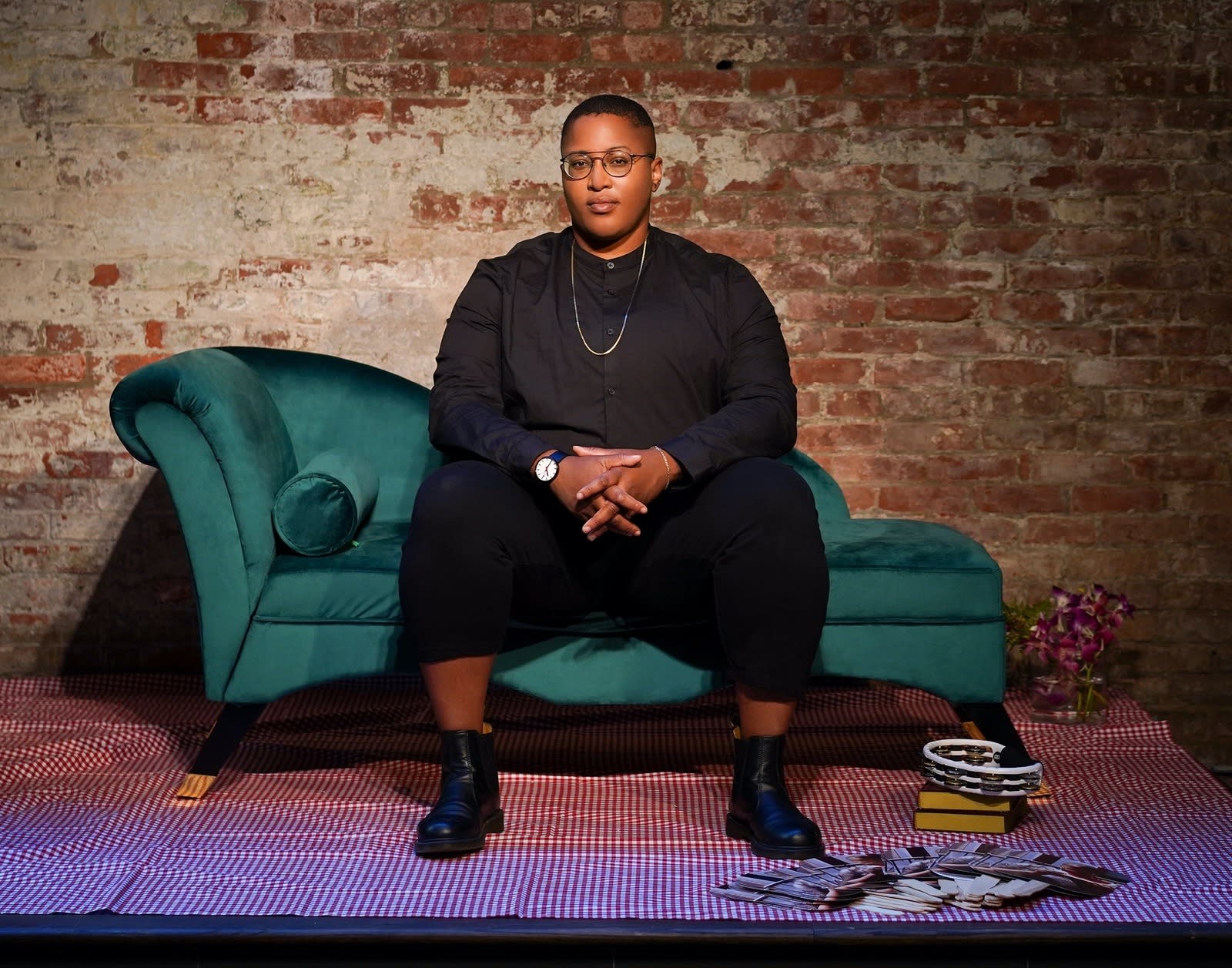 Photo of Sloan Leo wearing all black sitting upright on a teal chaise lounge in front of a brick wall.