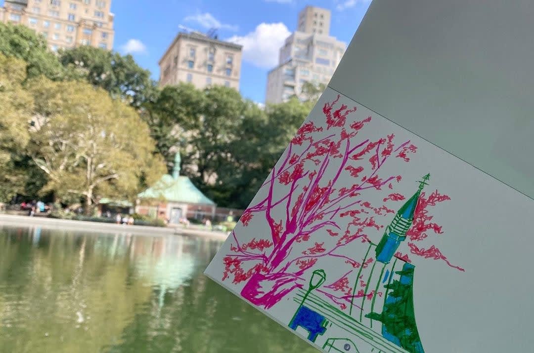 A sketch of a tree and building in a sketchbook is held in the air in front of a landscape in the distance.