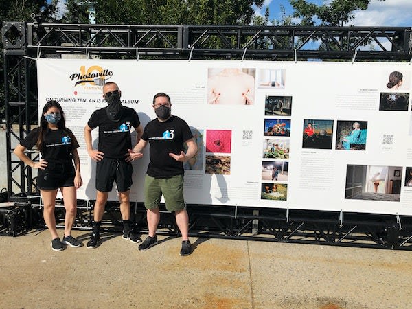 Three masked people stand outside in front of a large display/banner that has photos printed on it.