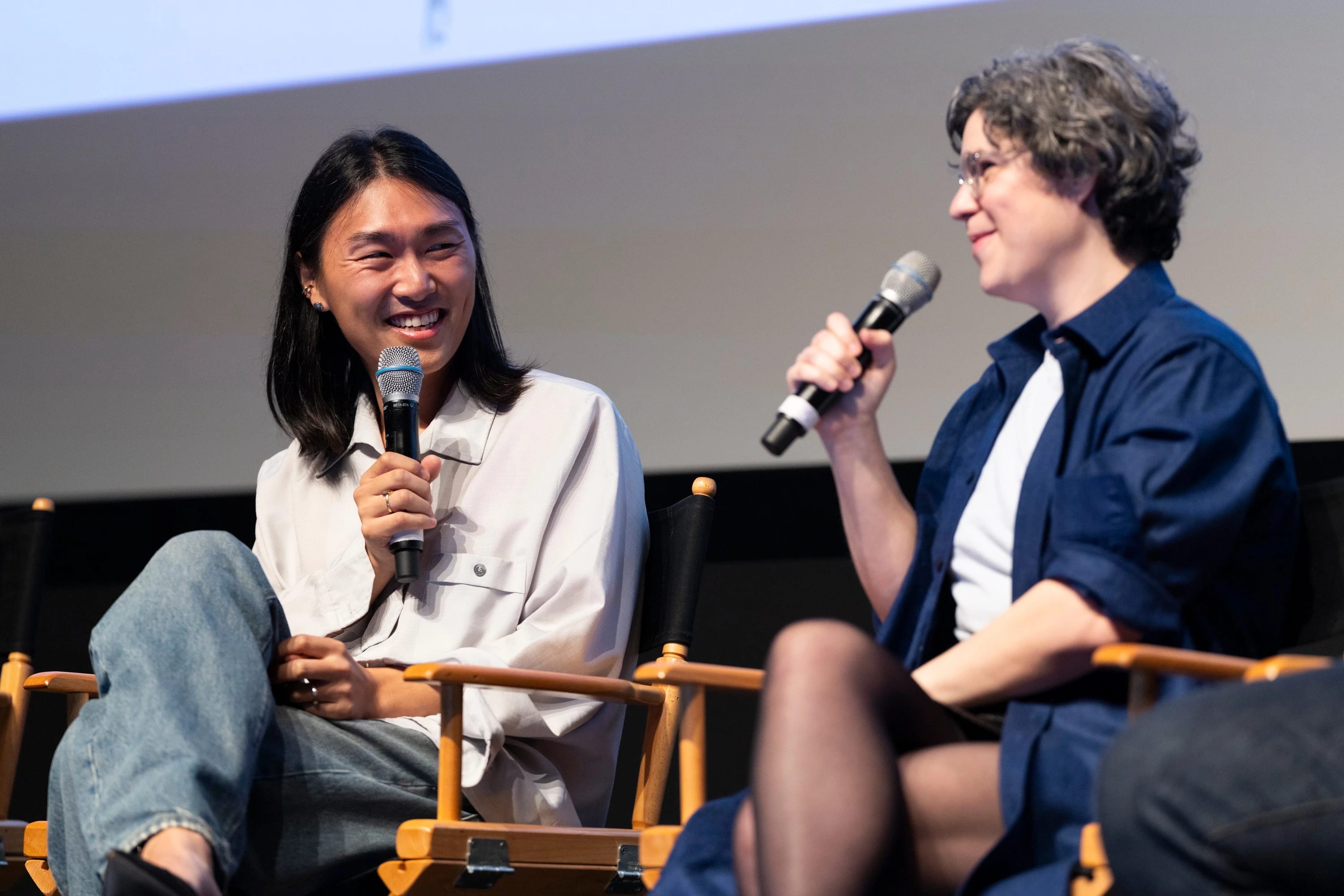 Two individuals sit cross-legged facing each other, each holding a microphone, engaged in conversation.