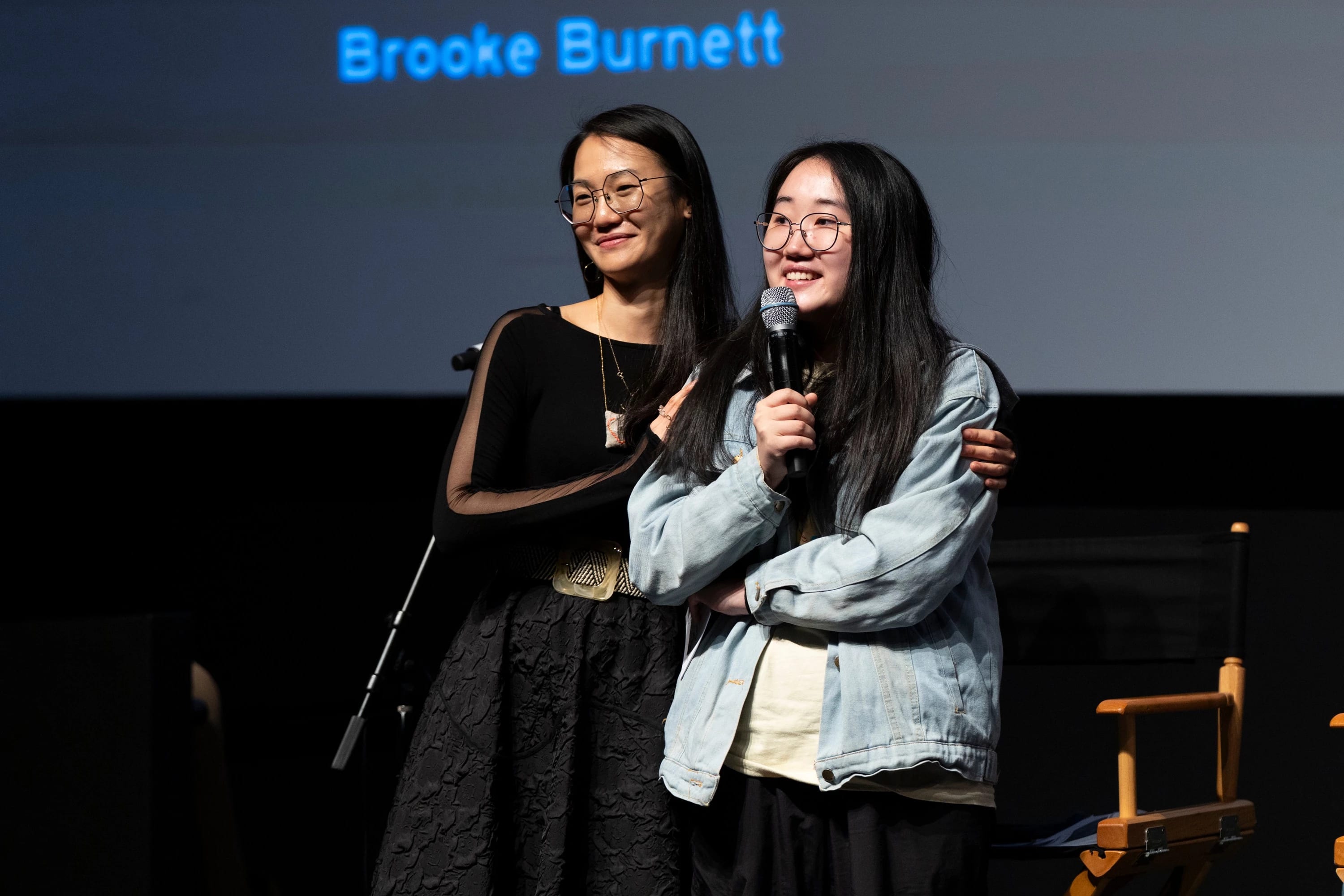 Two individuals stand close together on a stage, with one of them holding a microphone.