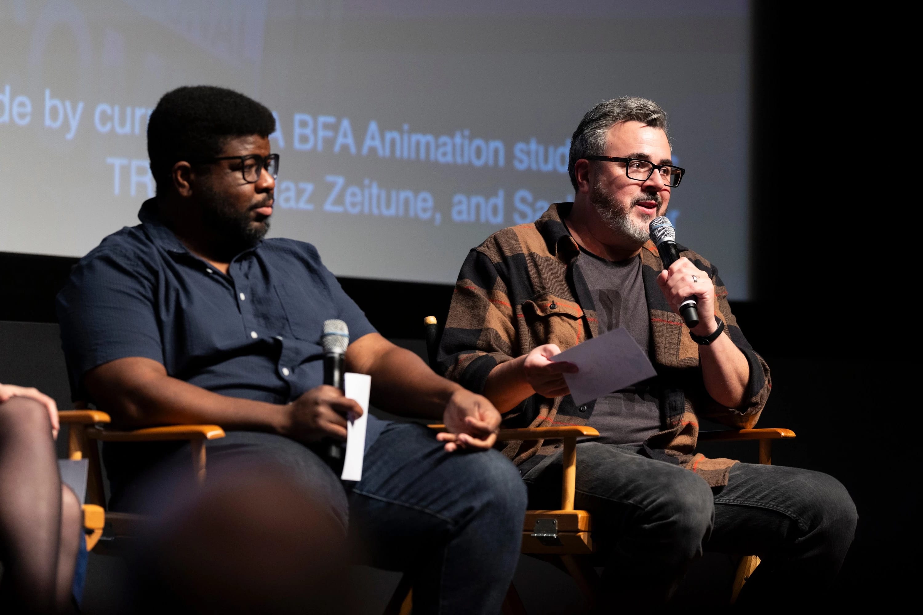 Two individuals are on a stage; one is holding a microphone and speaking.